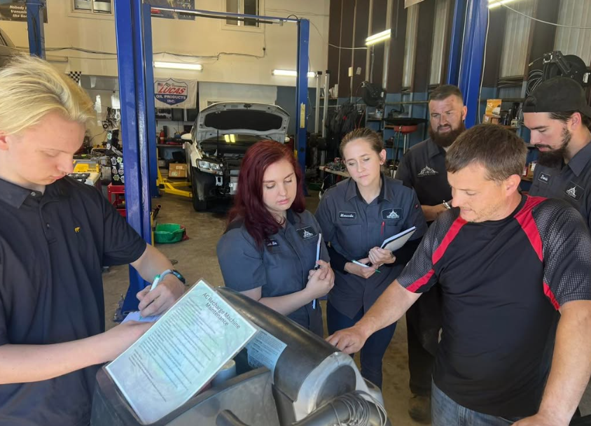 Group of people in a garage, some wearing mechanic uniforms, gathered around a service counter, discussing a vehicle repair.