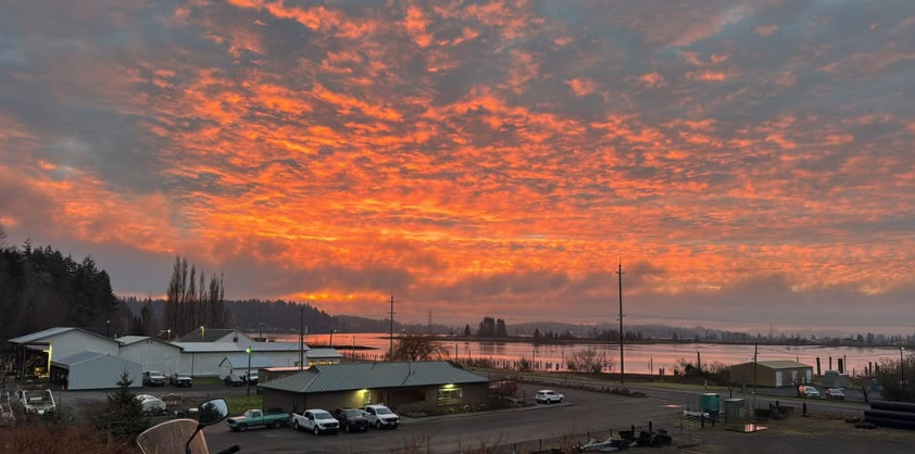 Sunset sky with orange and pink clouds over a harbor with boats and buildings, calm water reflecting the sky.