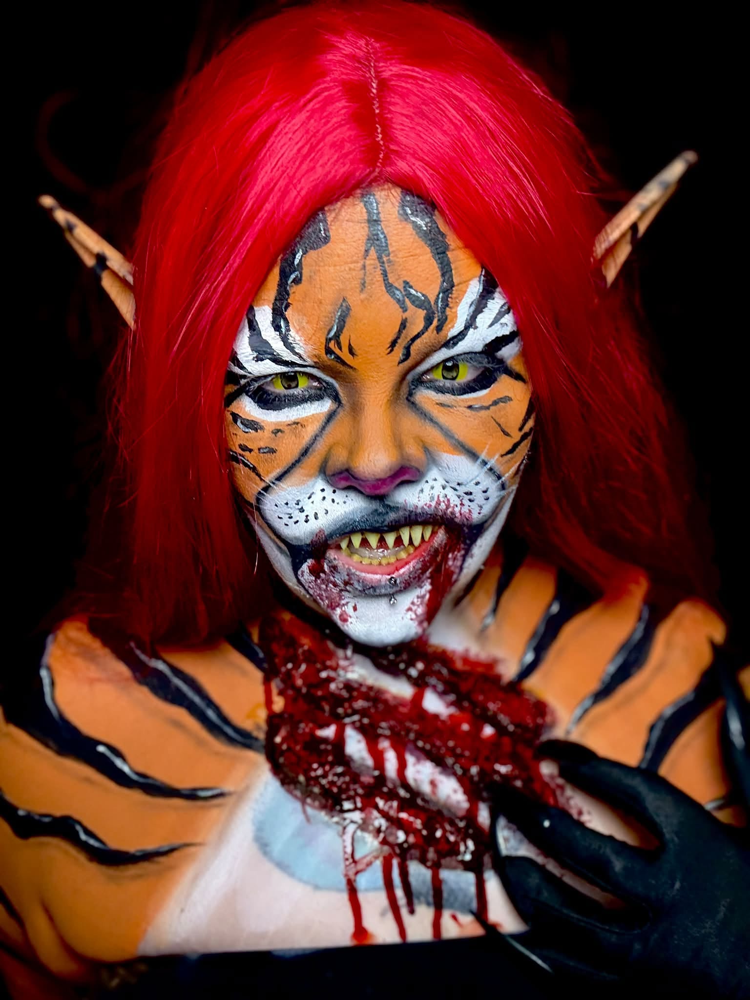 Person with tiger face paint, red hair, pointed ears, and fangs, holding a bloody prop, looking intensely at the camera.