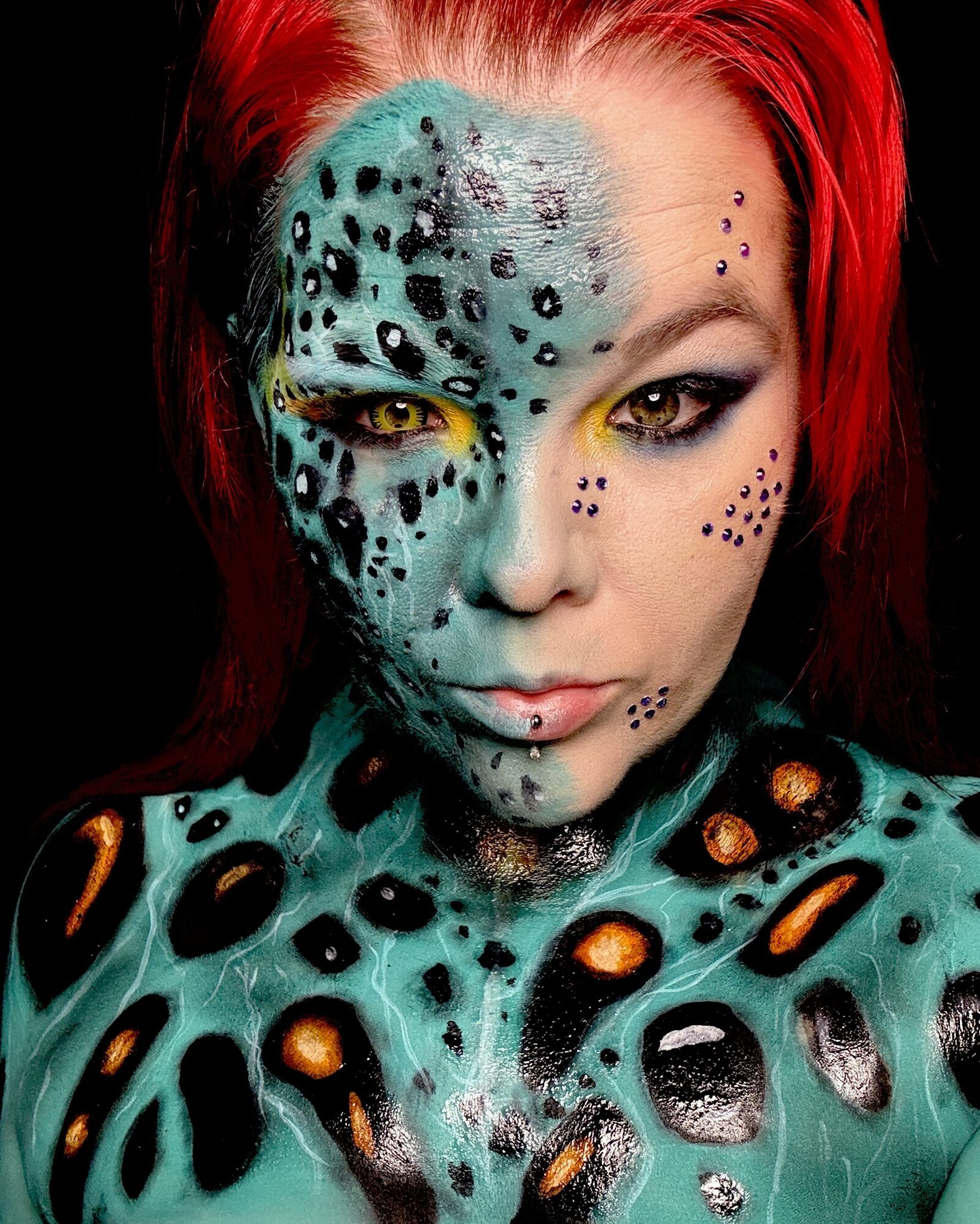 Woman with face and body painted as a leopard, with red hair and yellow eyes, looking directly at the camera.