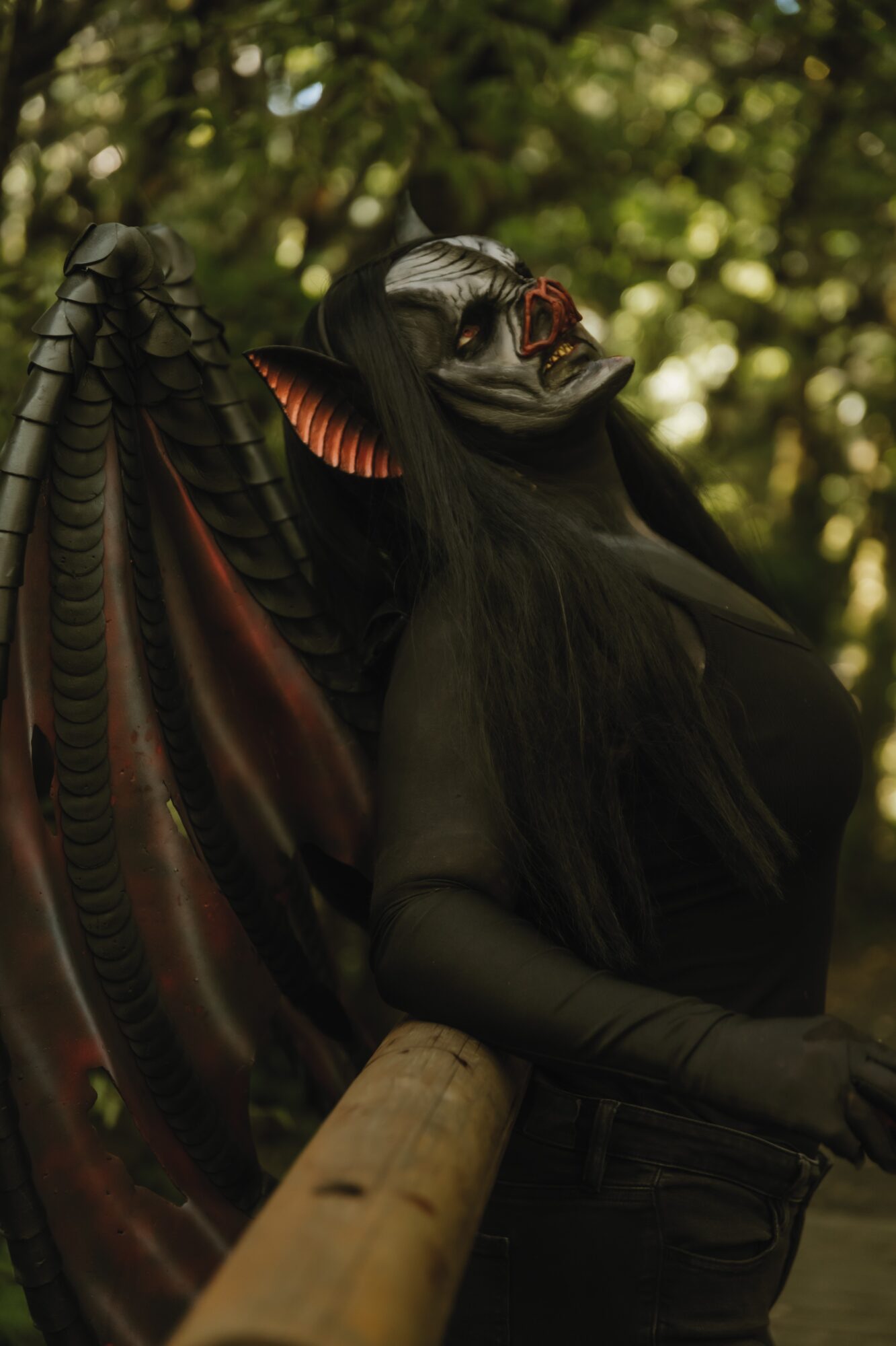 Person in elaborate costume with dark makeup and headpiece, outdoors with blurred green foliage background.