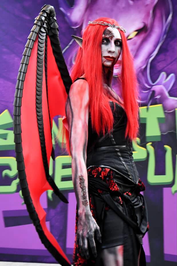 Person with red hair, pale skin, and elaborate costume, standing in front of a colorful background with purple and green elements.