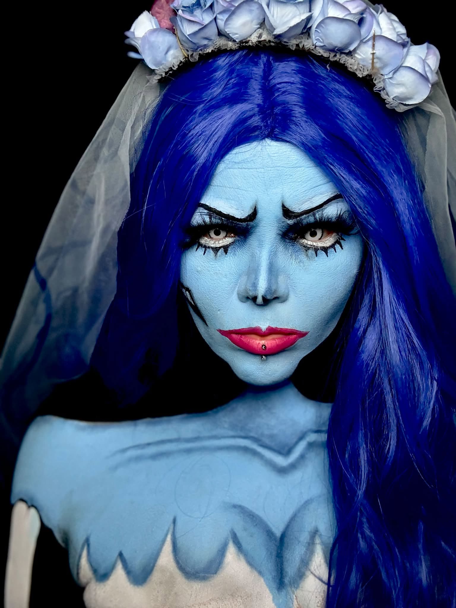 Person with blue skin, long blue hair, intense makeup, and a floral headpiece, wearing a costume with jagged edges.