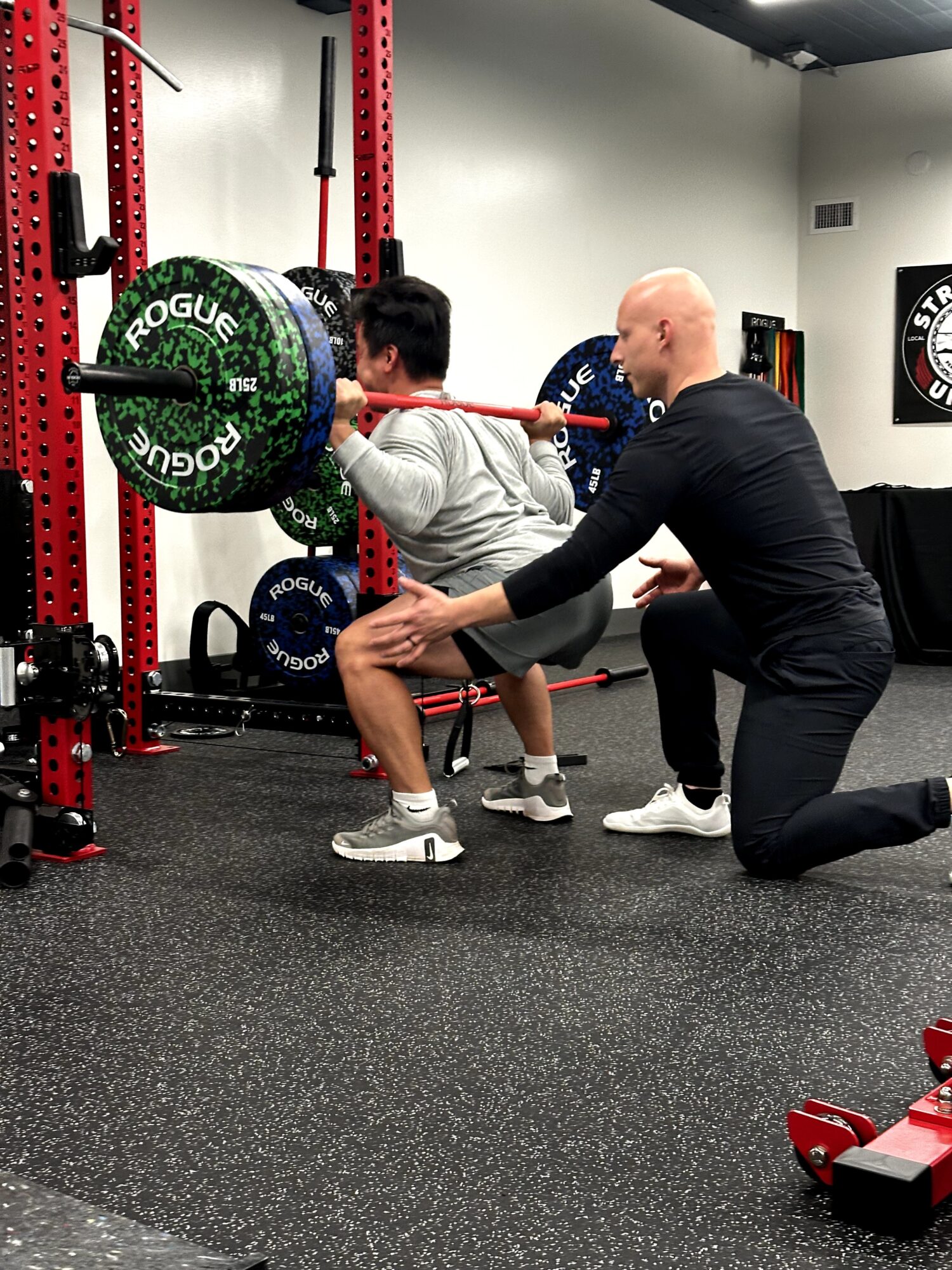 Person performing squat with barbell on shoulders, trainer assisting in gym setting, weight plates and equipment visible