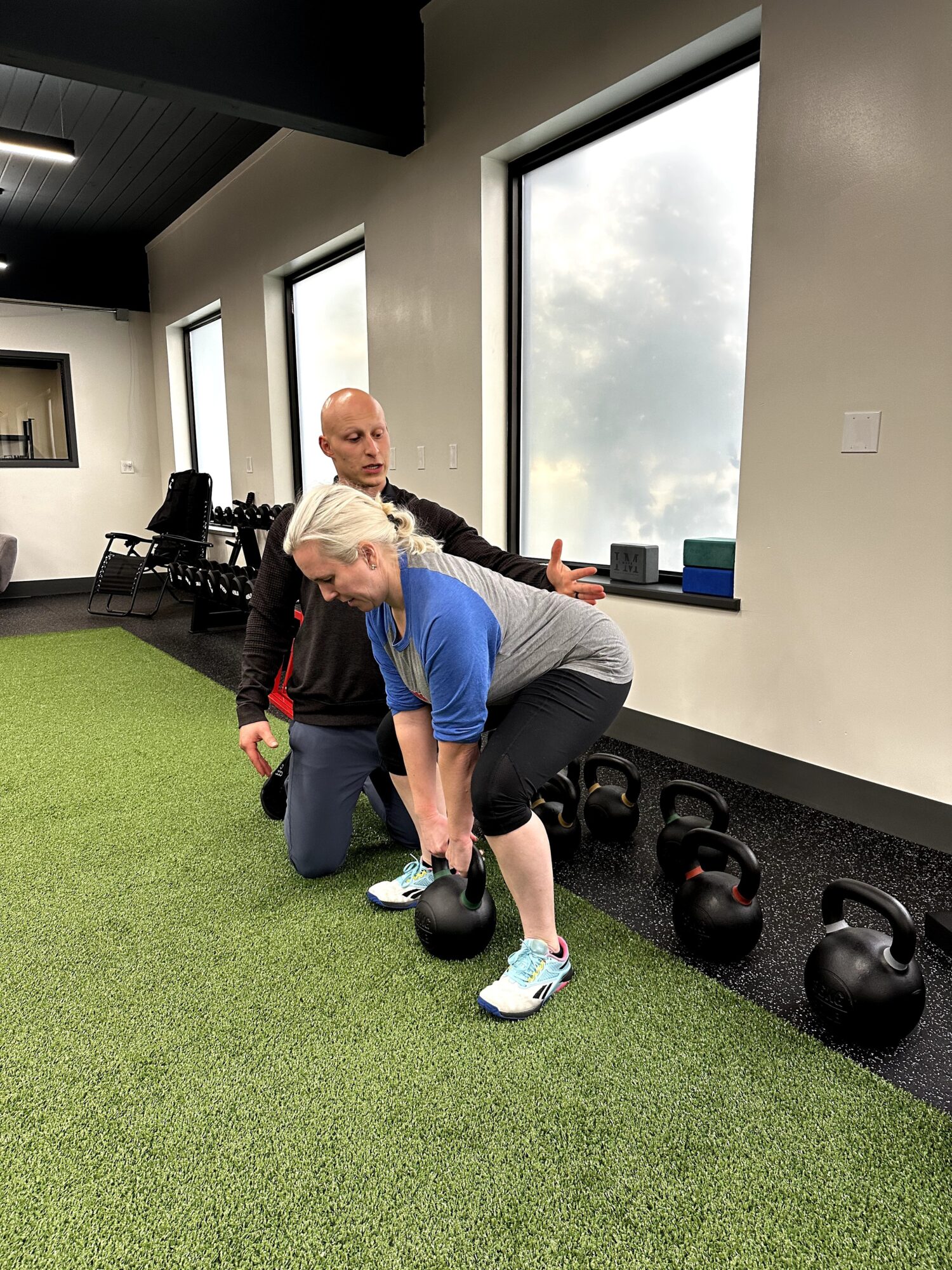 Woman lifting kettlebell with assistance from trainer in gym, large windows in background, green turf floor.