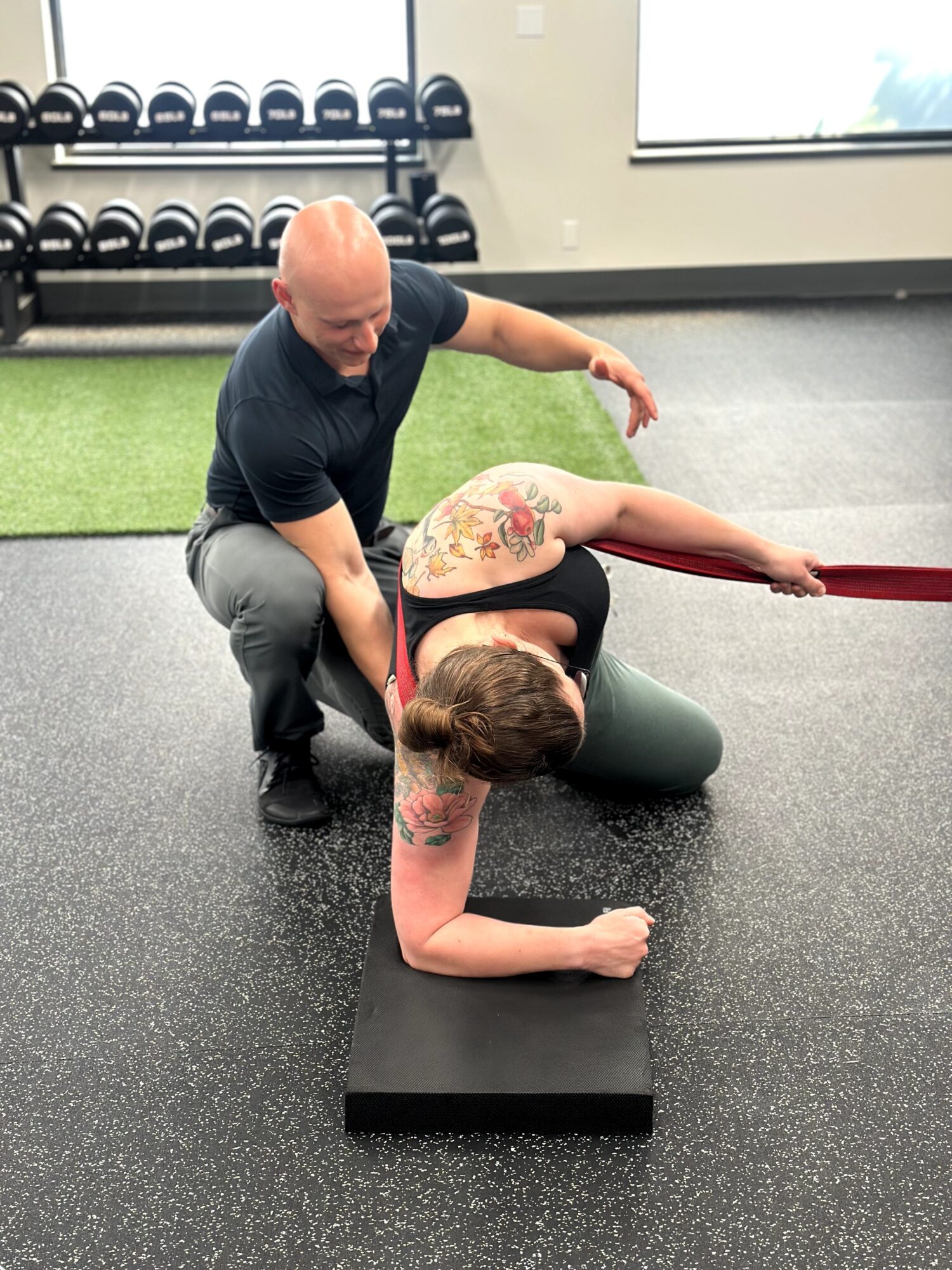Trainer assisting a woman with a tattoo in a stretching exercise on a mat in a gym, with weights and window in background.