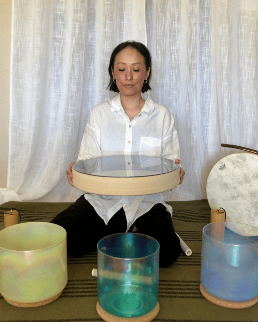 Person sitting cross-legged holding a large round drum, with three smaller drums in front, against a light curtain background.