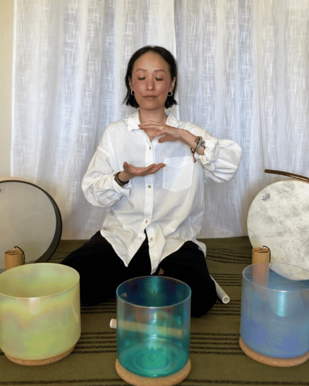 Woman sitting cross-legged with eyes closed, hands in meditation, three colorful glass bowls in front, white curtain background.