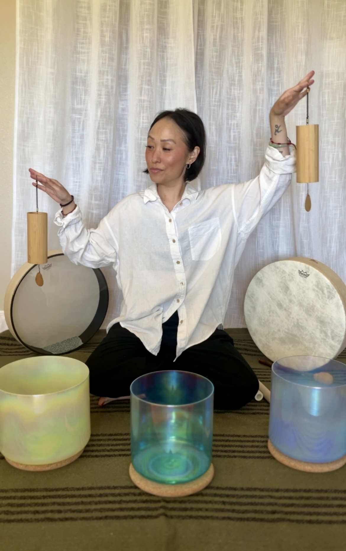 Woman sitting cross-legged with arms raised, surrounded by colorful glass bowls, in front of a light curtain backdrop.
