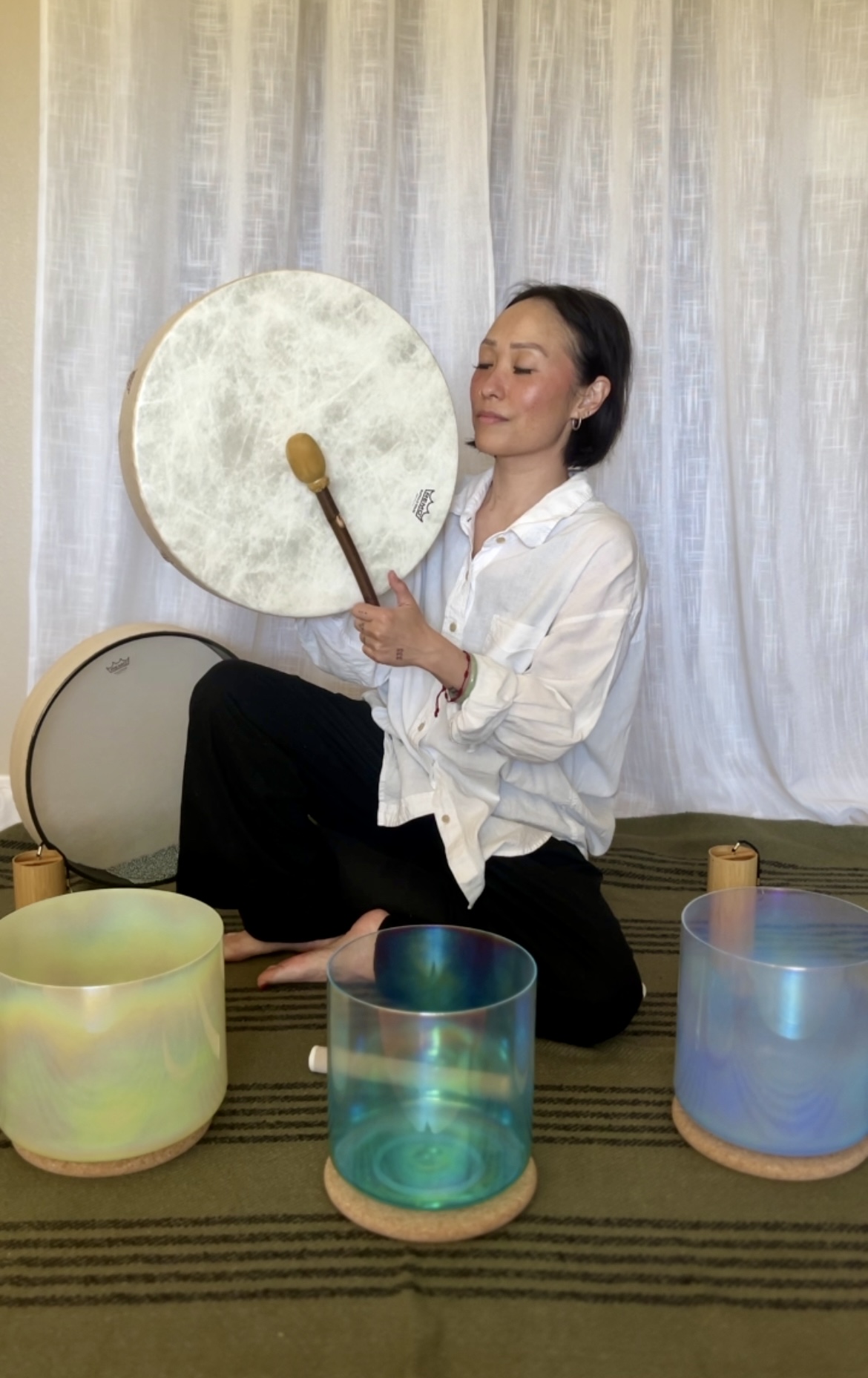 Woman sitting cross-legged, holding a large drum and a drumstick, with colorful drums in front, against a curtain background.