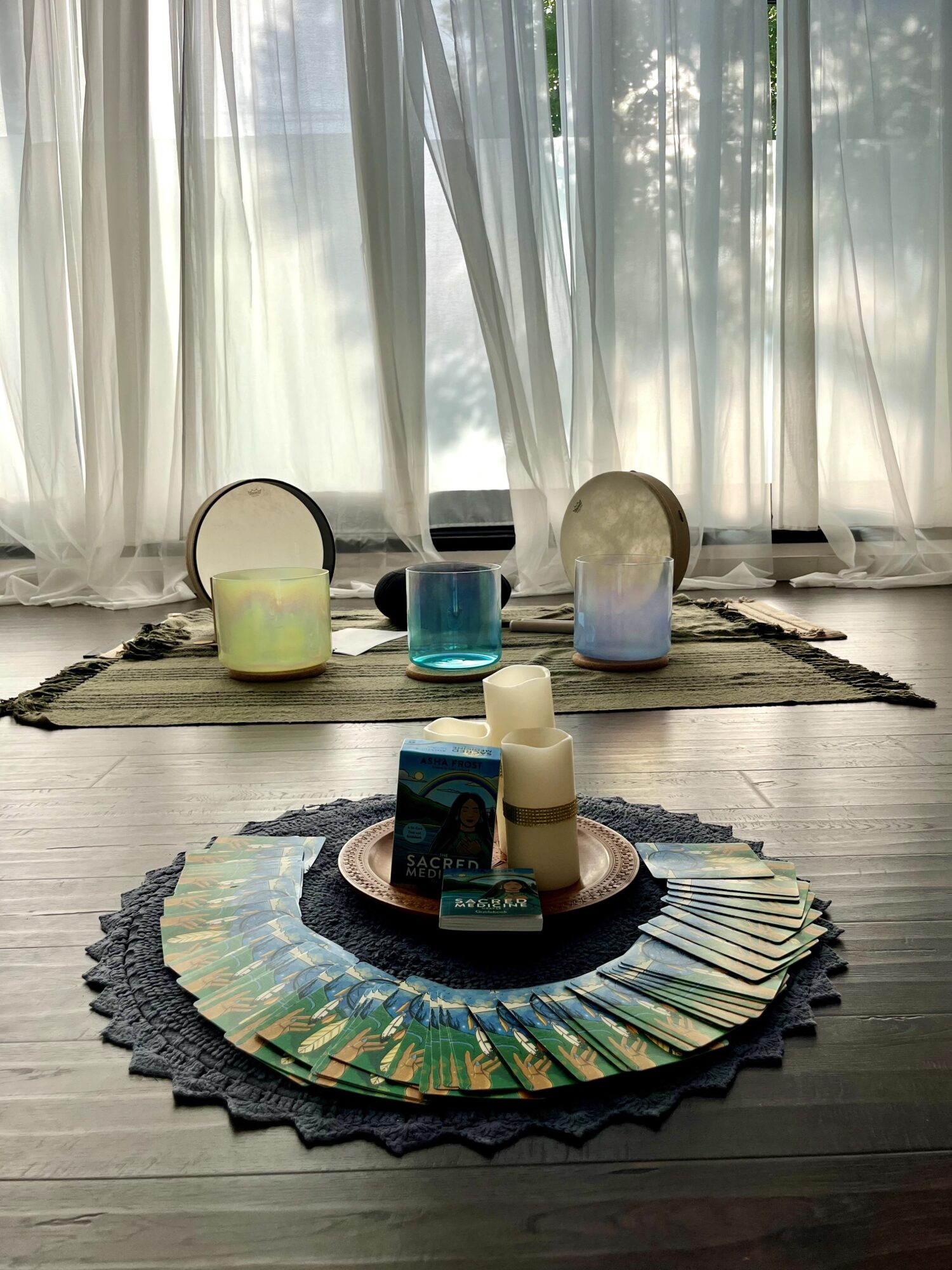 Decorative candles and objects arranged on a table in front of a window with sheer curtains, sunlight filtering through.