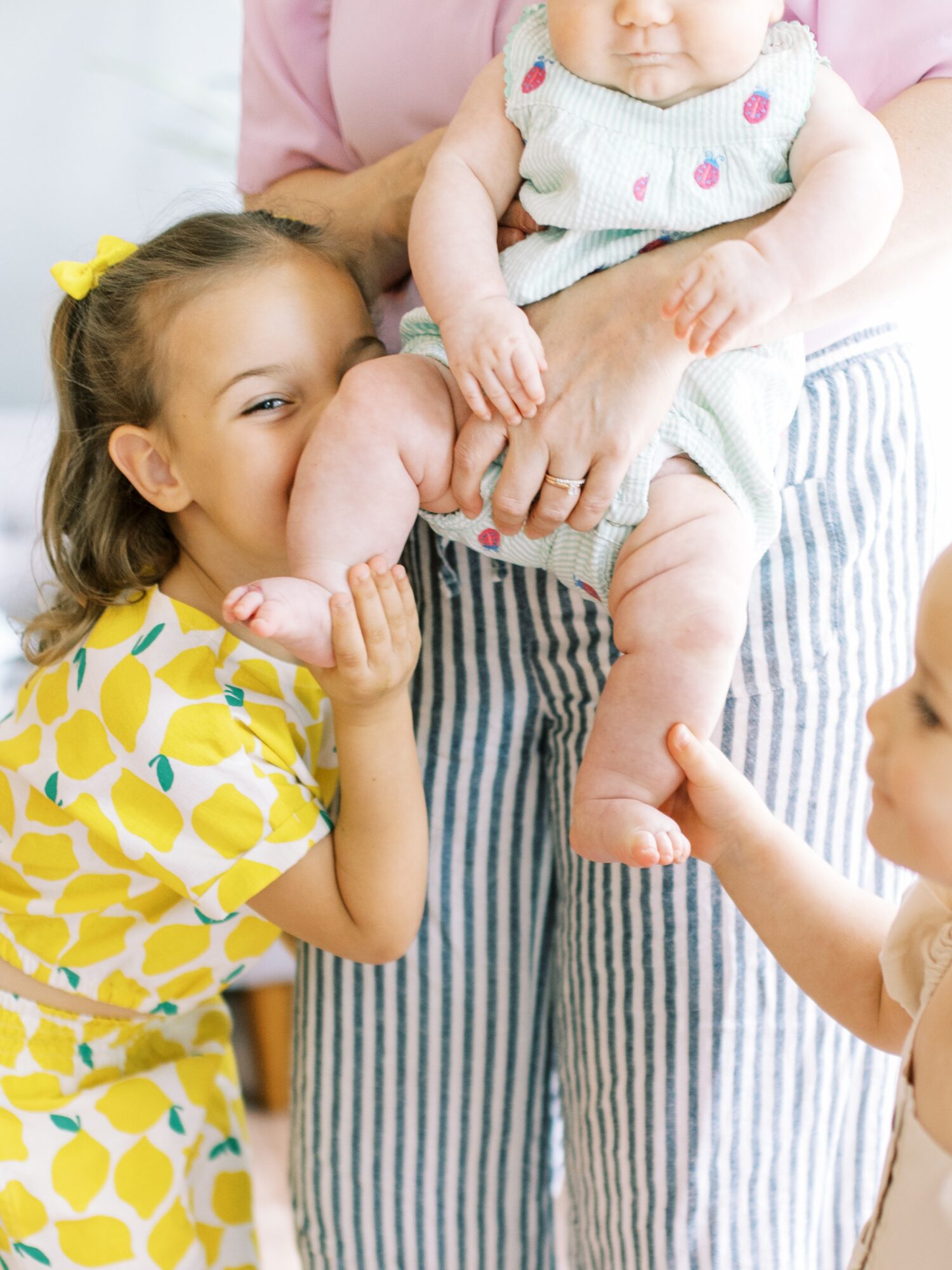 Two children and an adult holding a baby, with the girl smiling and touching the baby's foot, in a bright room.