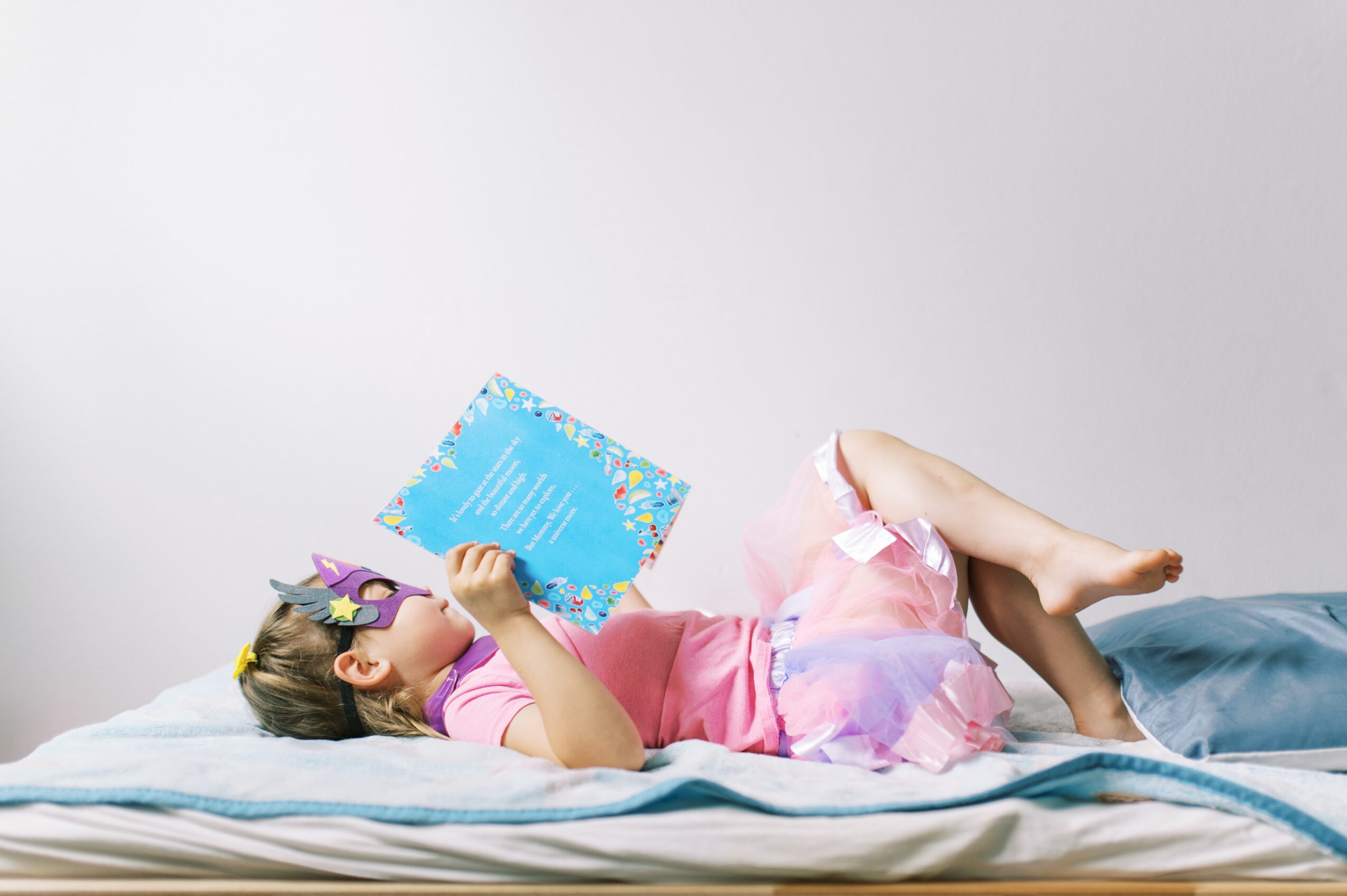 Child lying on bed reading a colorful book, wearing pink pajamas and glasses, with a plain light background.