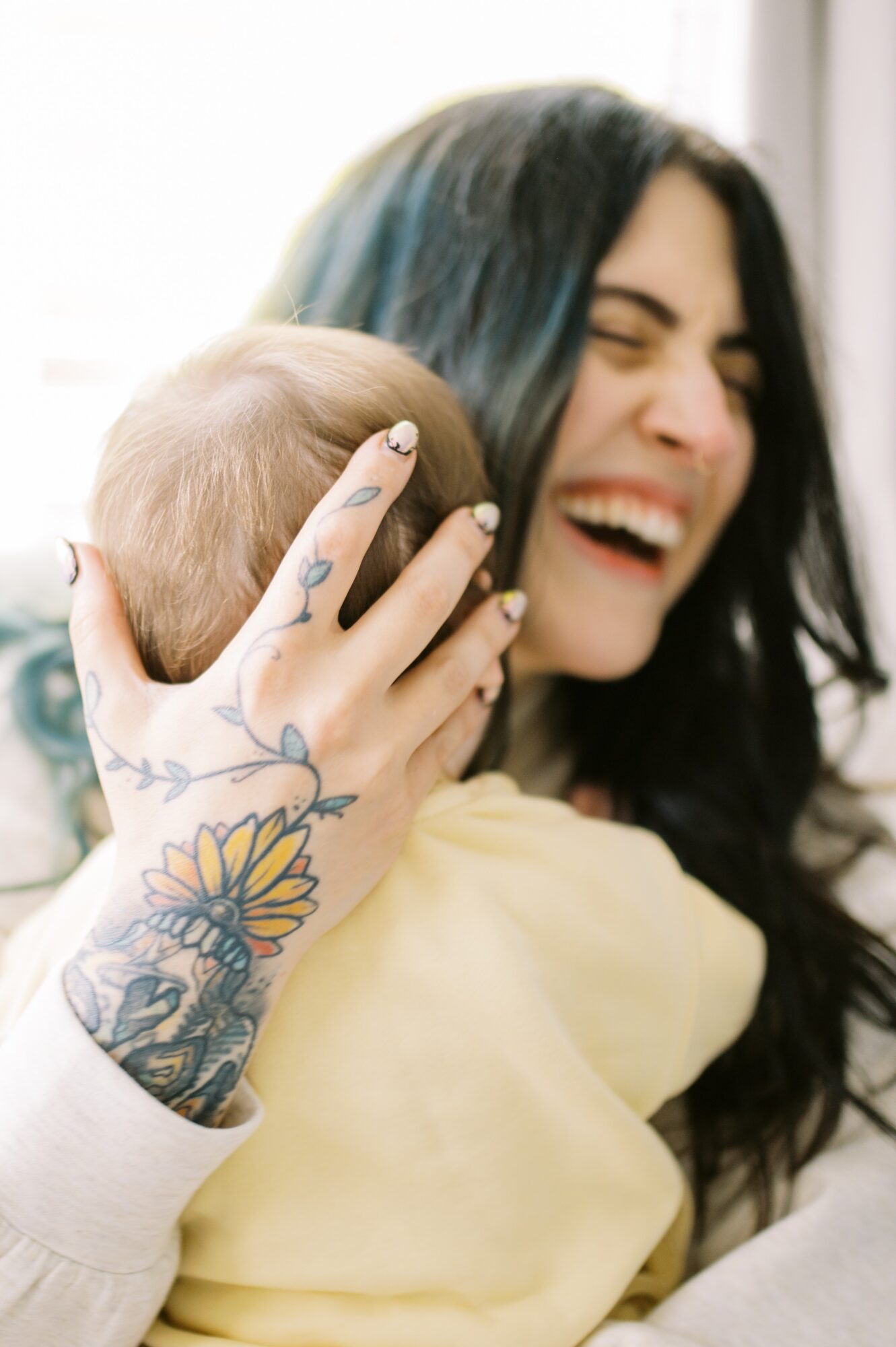 Woman with long dark hair laughing, holding a child with a tattooed hand, both smiling, indoors with light background.