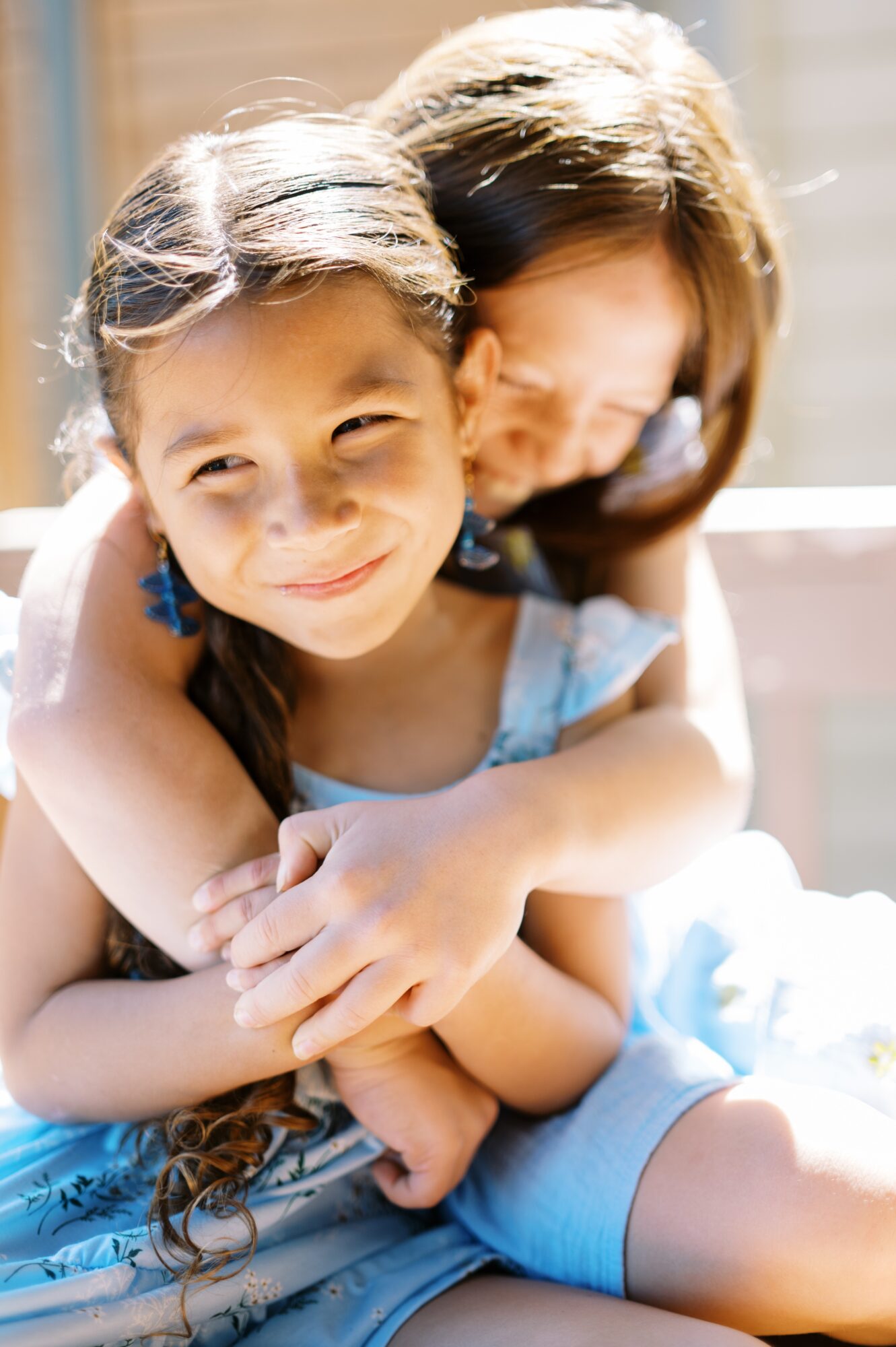 Child smiling with an adult hugging her from behind outdoors, sunlight shining, both appearing happy.