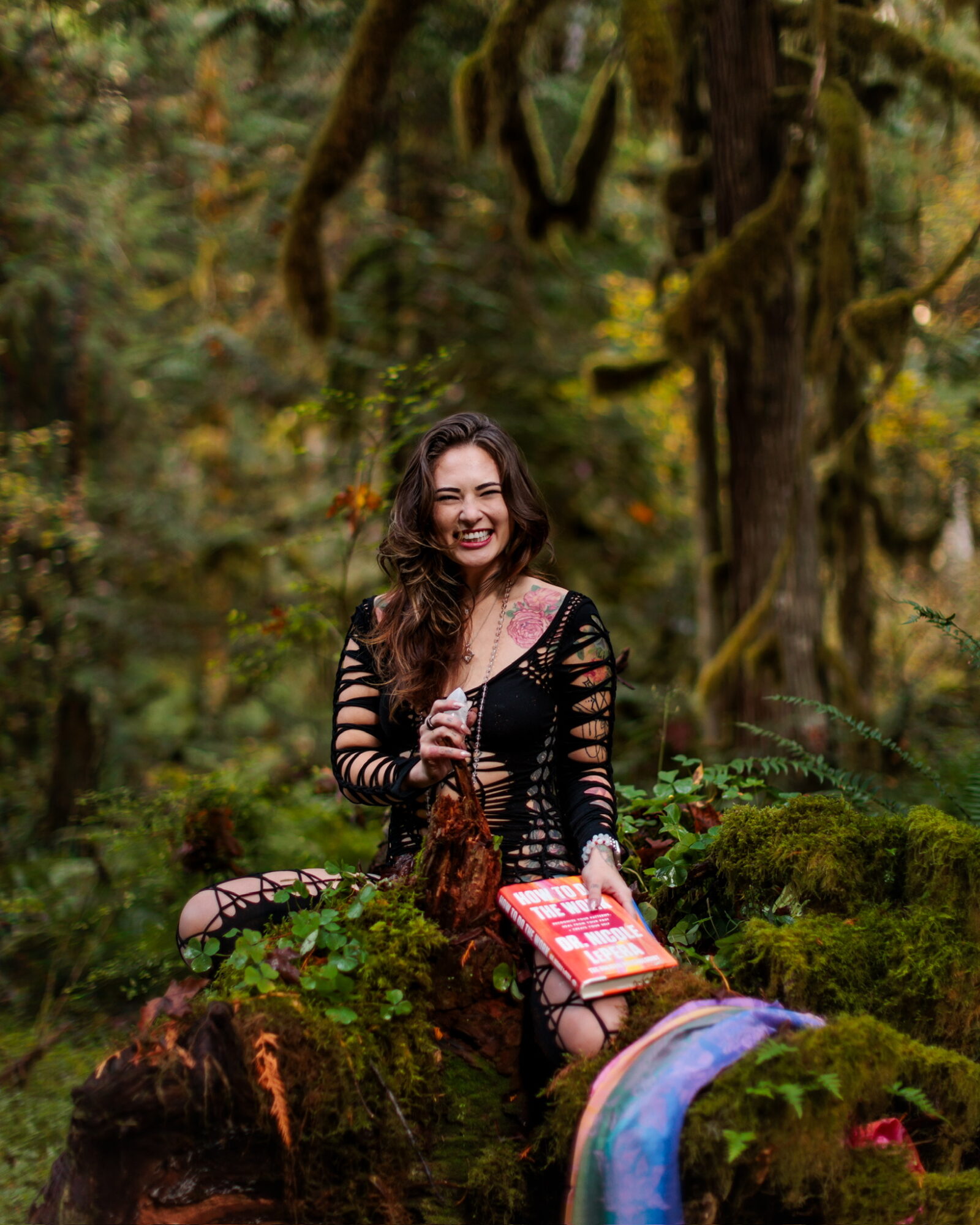 Young woman with long dark hair smiling in a forest, sitting on a mossy log holding a book, surrounded by trees and greenery.