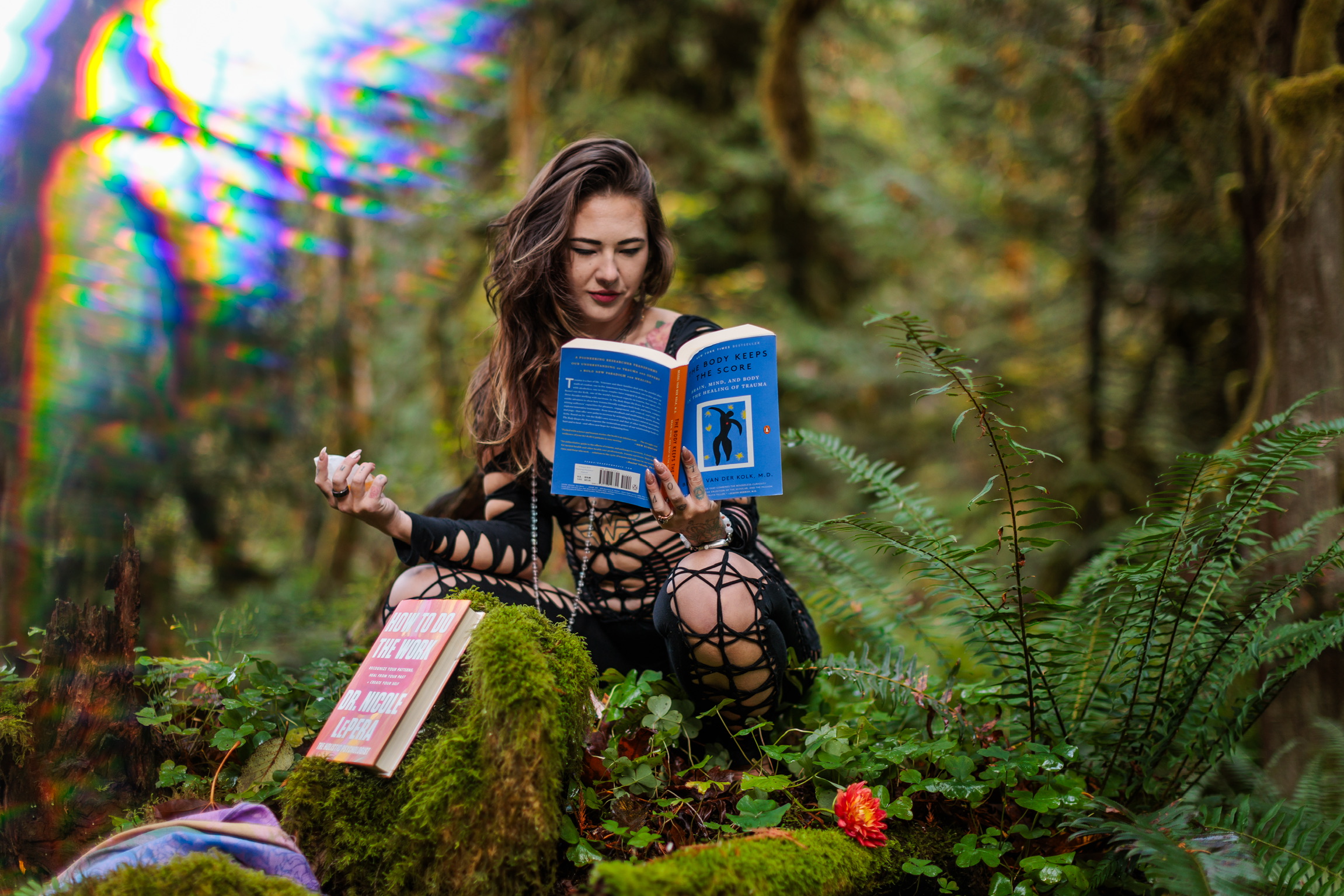 Young woman reading a book while sitting on a moss-covered log in a forest, surrounded by greenery and trees.
