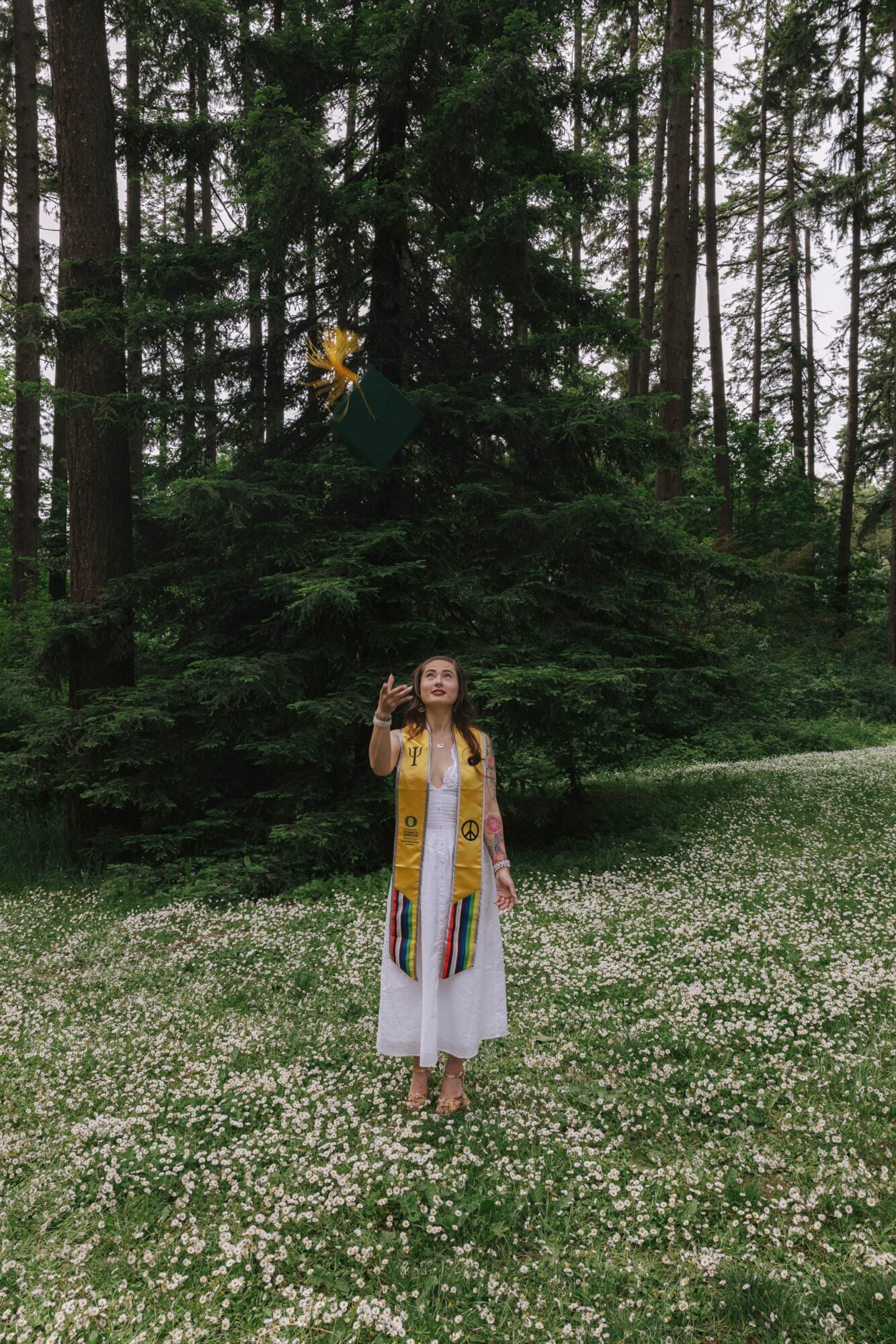 Woman in white dress and colorful scarf standing in a forest clearing with trees and flowers, holding a yellow flower.