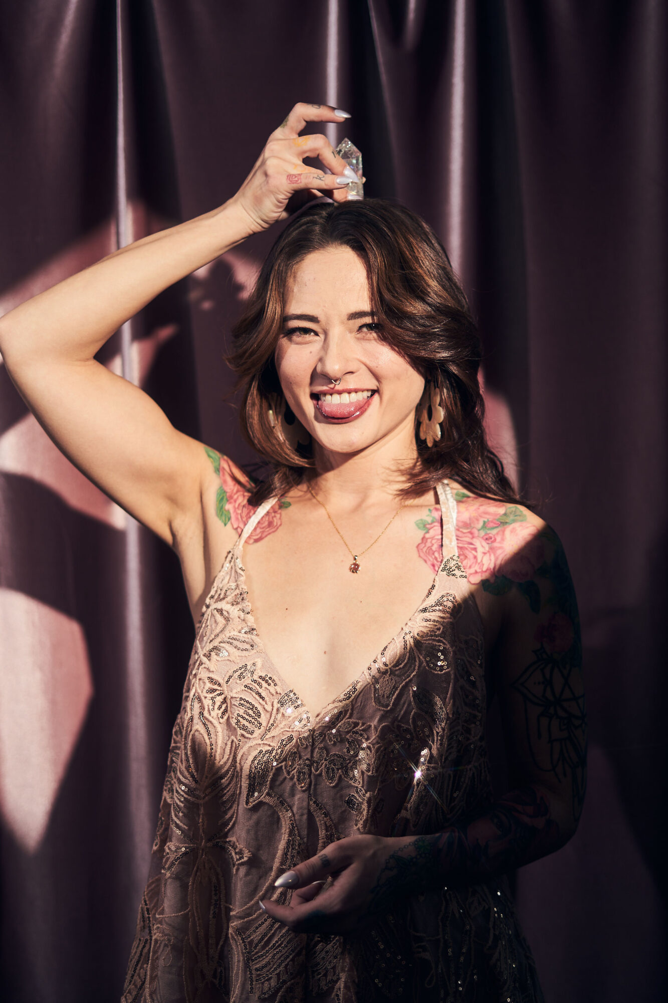 Woman smiling, holding a glass above her head, standing in front of dark curtains, wearing a lace dress.