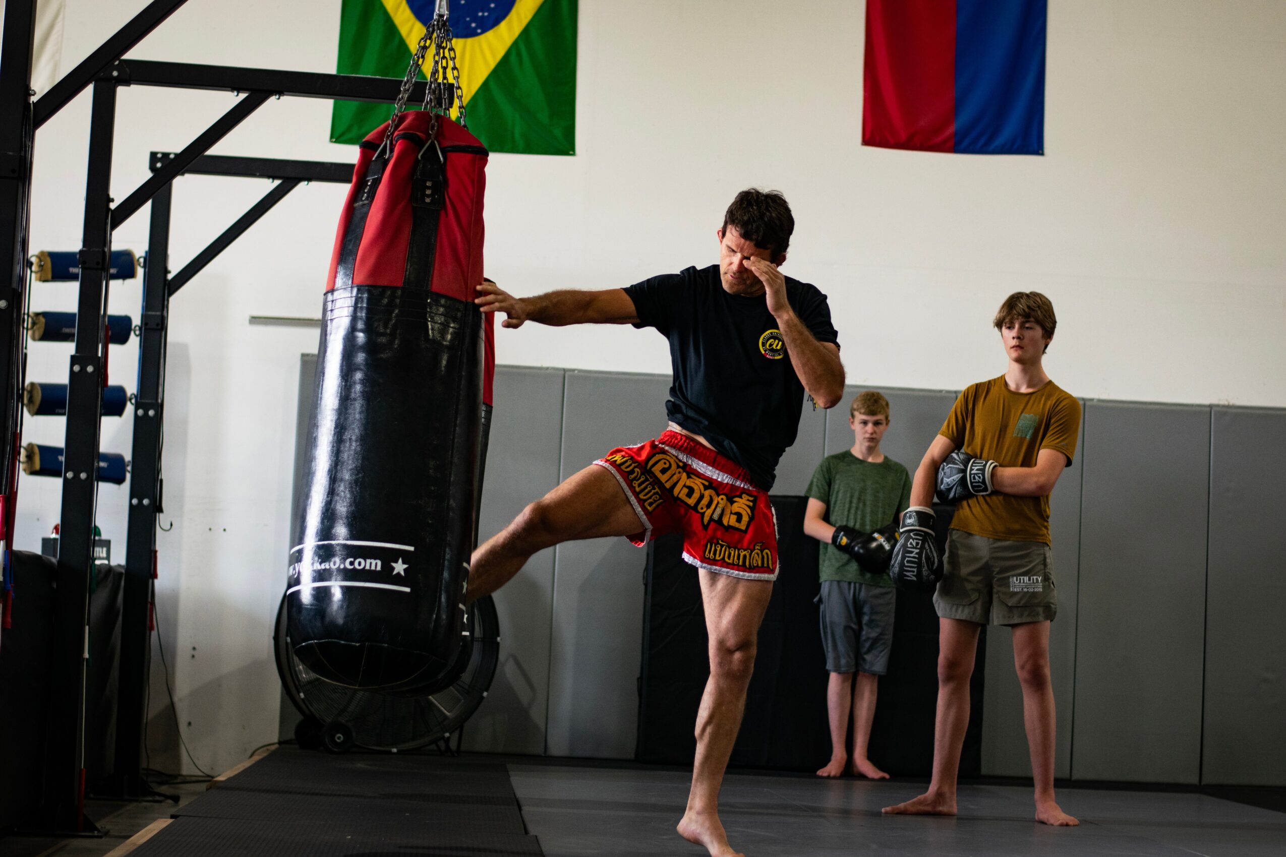 Person practicing martial arts kicks a hanging punching bag in a gym with flags on the wall, two children watch.
