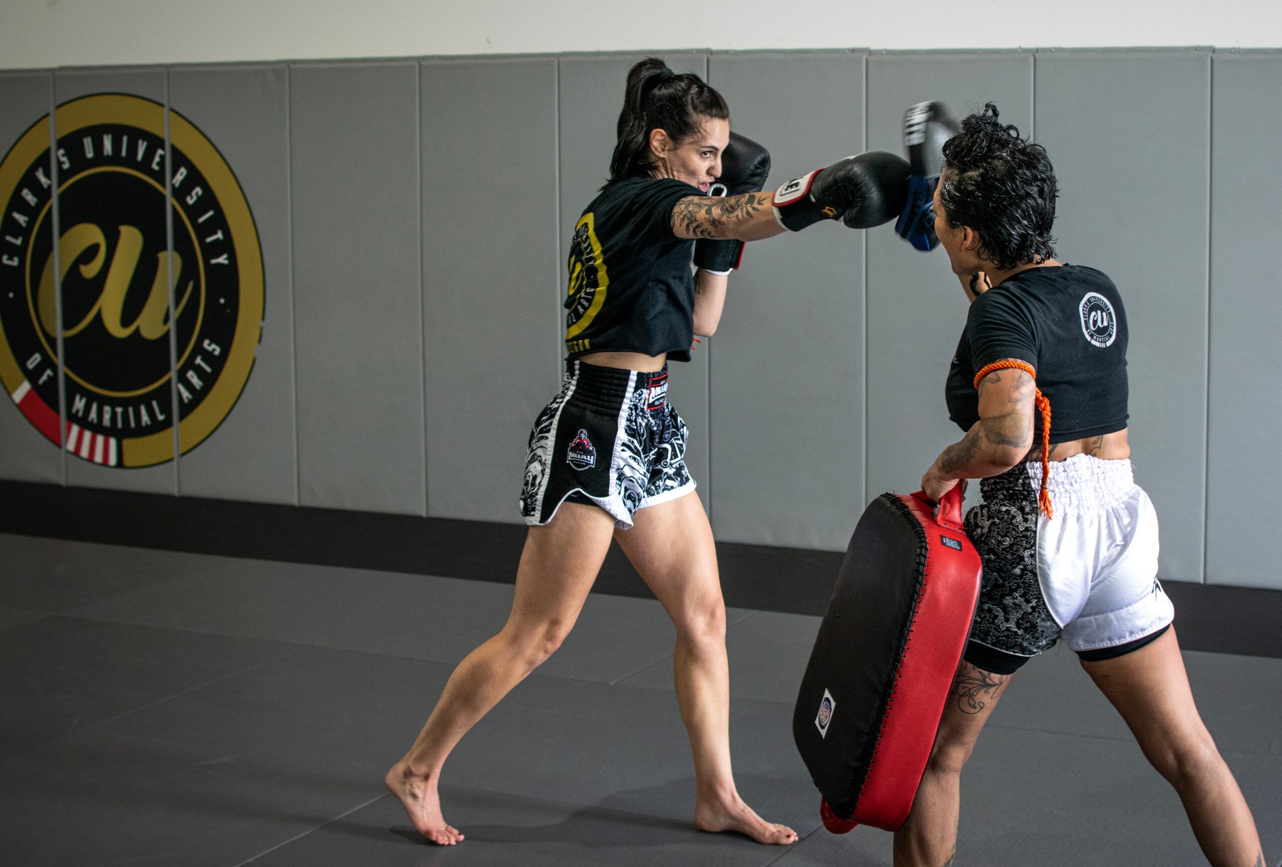 Two women practicing martial arts in a gym, one throwing a punch, the other holding a shield pad.