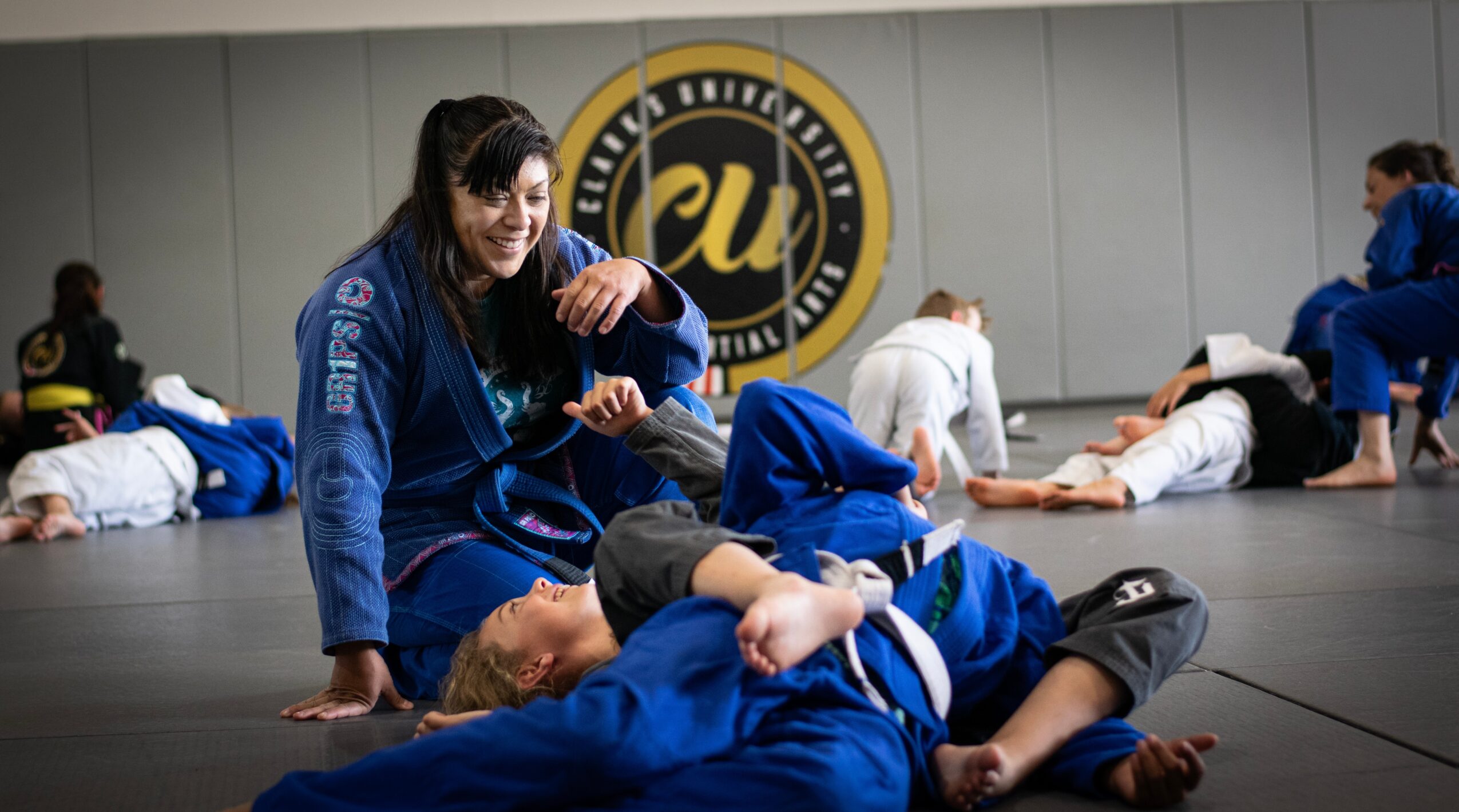 Brazilian Jiu-Jitsu training session with people practicing techniques on mats, some lying down, in a gym with a logo on the wall.