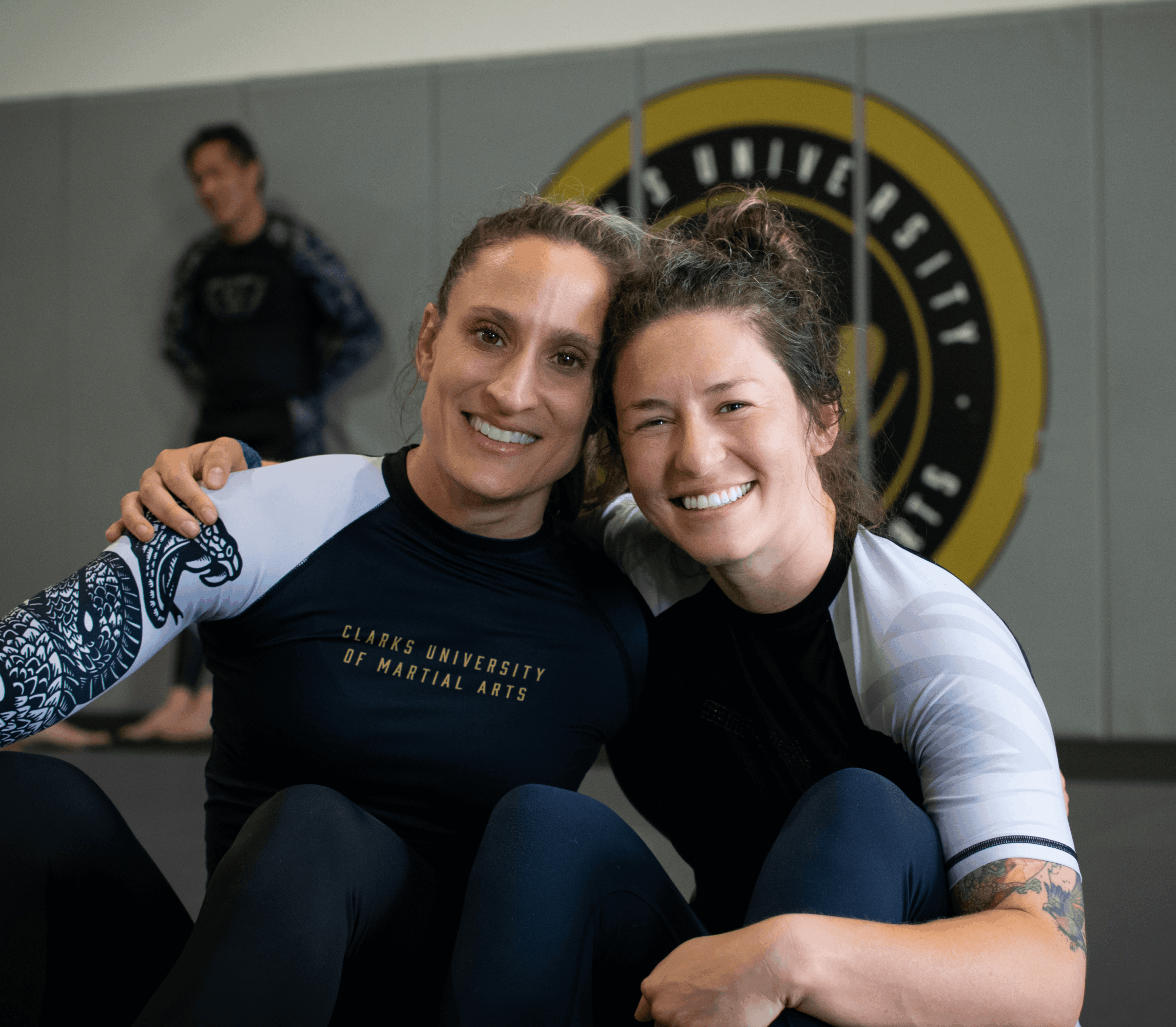 Two women smiling and hugging in a gym, with a logo on the wall behind them.