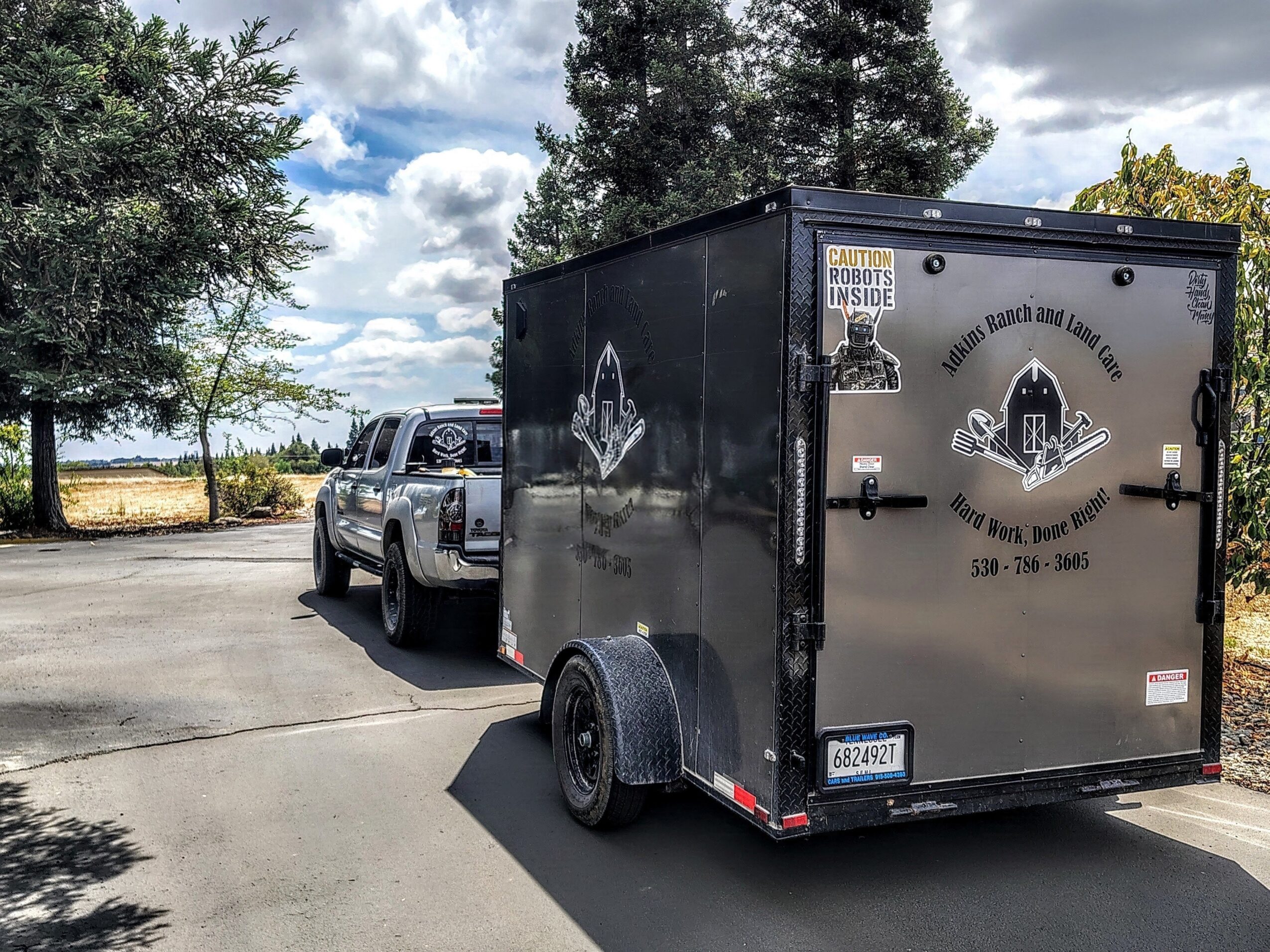 Black enclosed trailer attached to a pickup truck parked on a paved road with trees and cloudy sky in background.