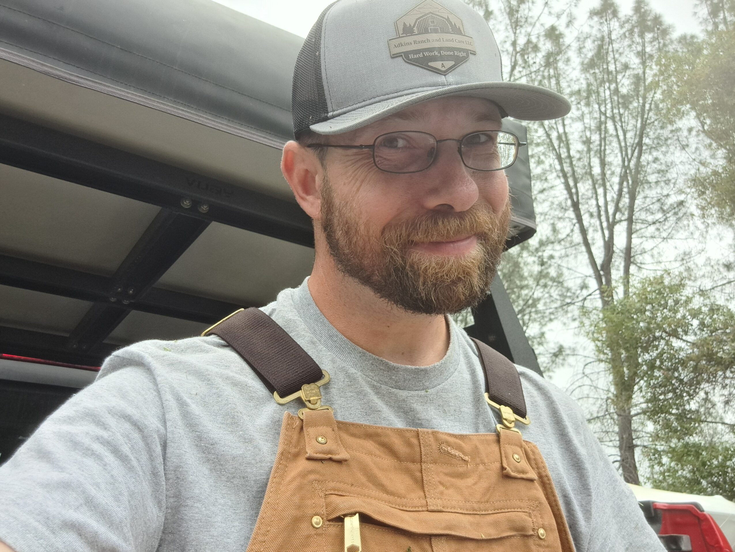 Man with glasses and beard wearing a gray cap and gray shirt, smiling outdoors with trees in background.