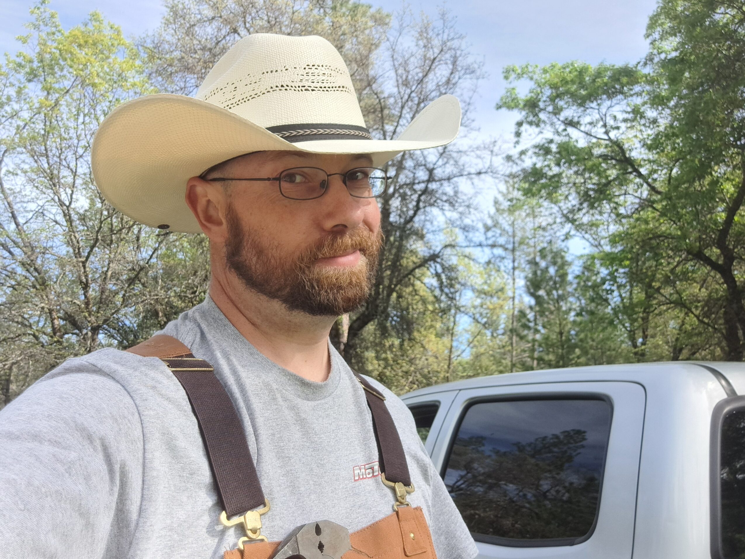 Man wearing a cowboy hat and glasses outdoors, with trees and a vehicle in the background.