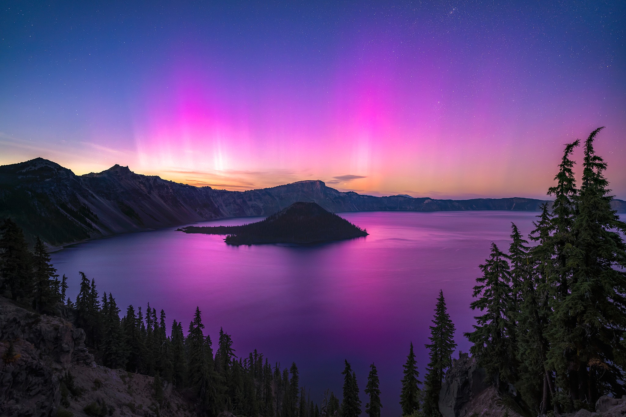 Night sky with colorful aurora over a lake, surrounded by mountains and trees, with reflections on the water.