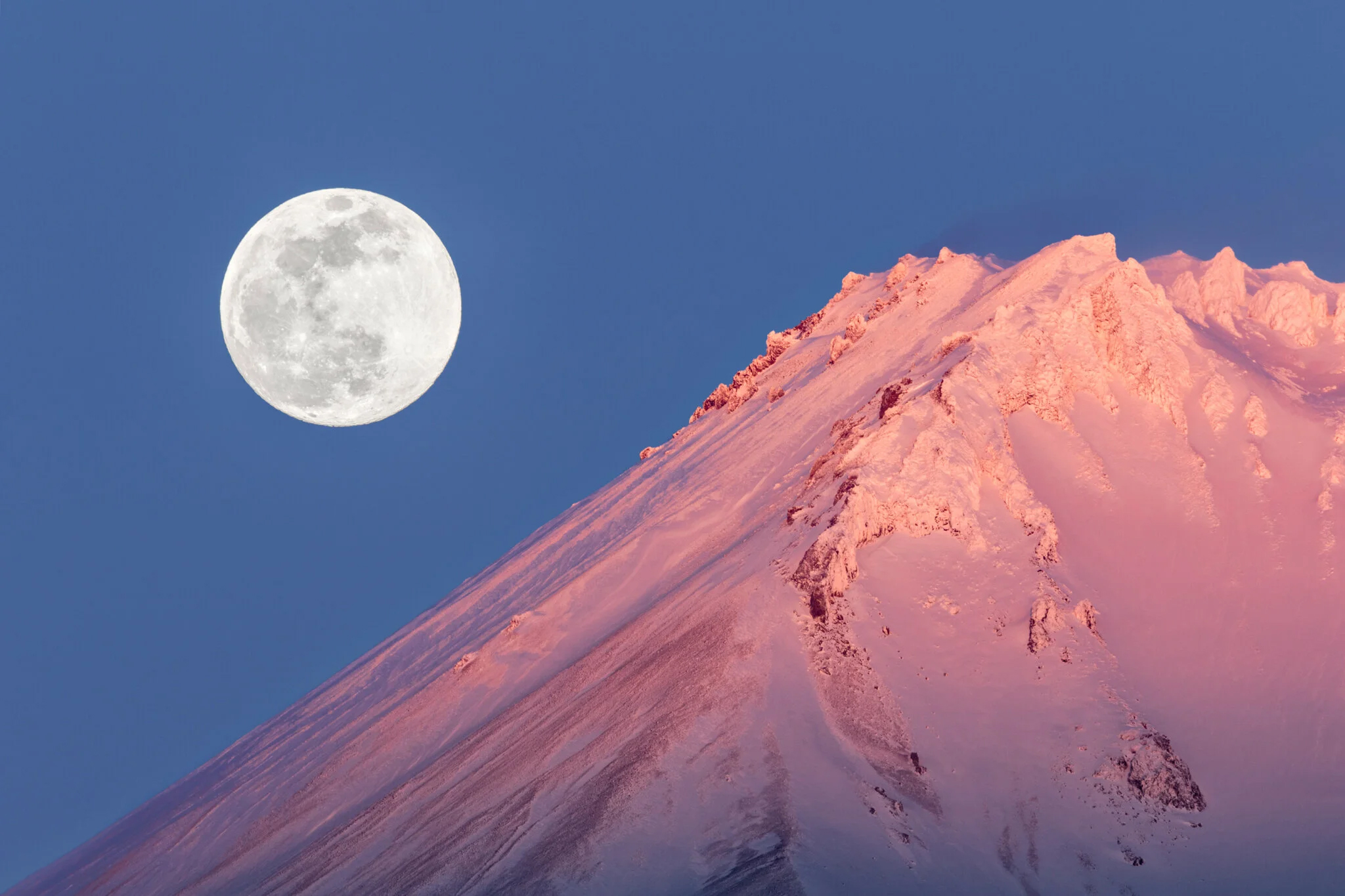 Full moon in a clear sky above a snow-capped mountain with pinkish hues, possibly during sunrise or sunset.