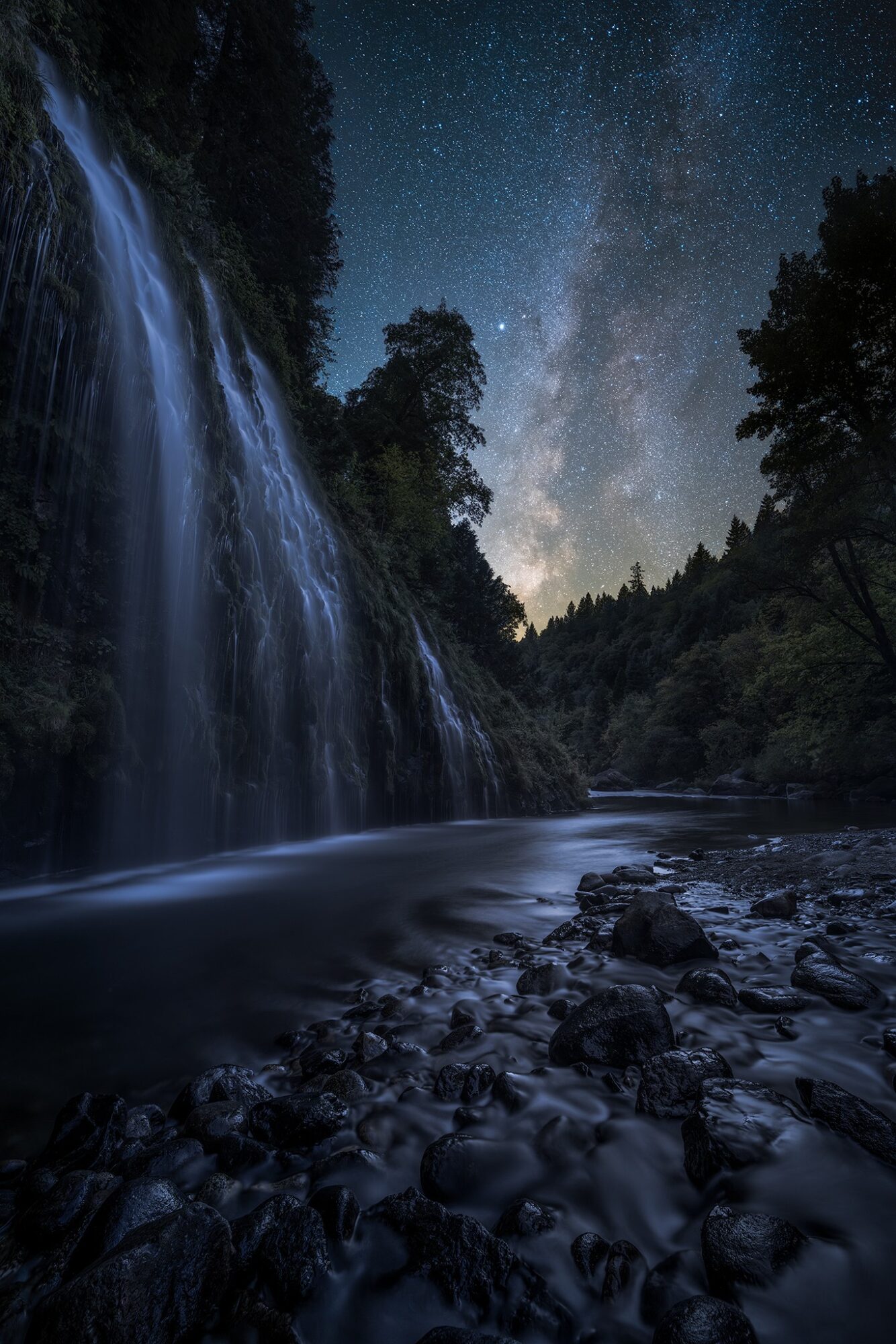 Nighttime scene of a waterfall flowing into a river, with a starry sky and trees in the background.