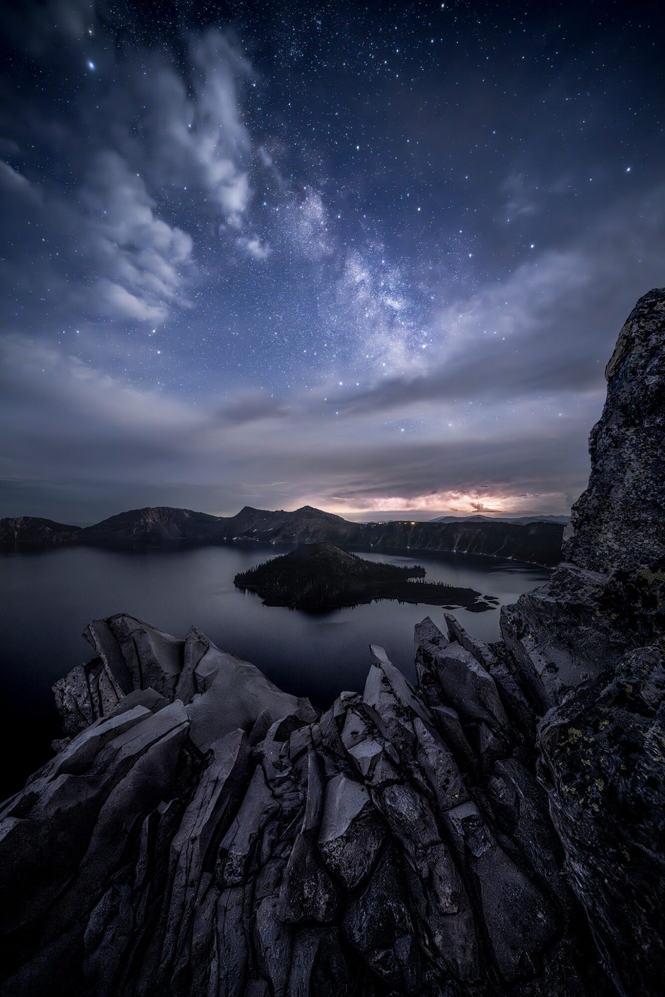 Night sky with stars and Milky Way over a rocky landscape and calm water, with clouds and a distant horizon.
