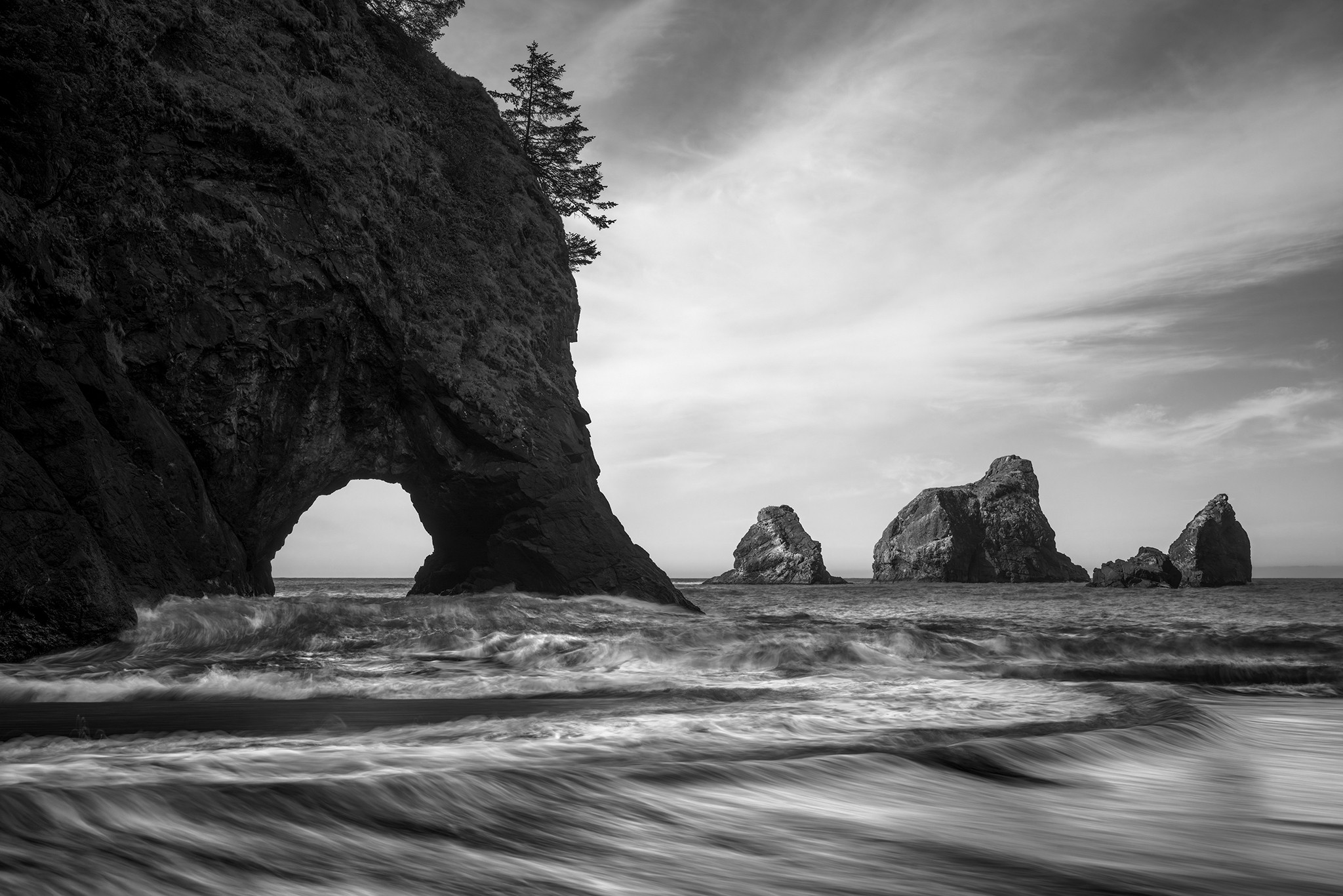 Cliff with natural arch on the left, ocean waves, and rock formations in the distance under cloudy sky.