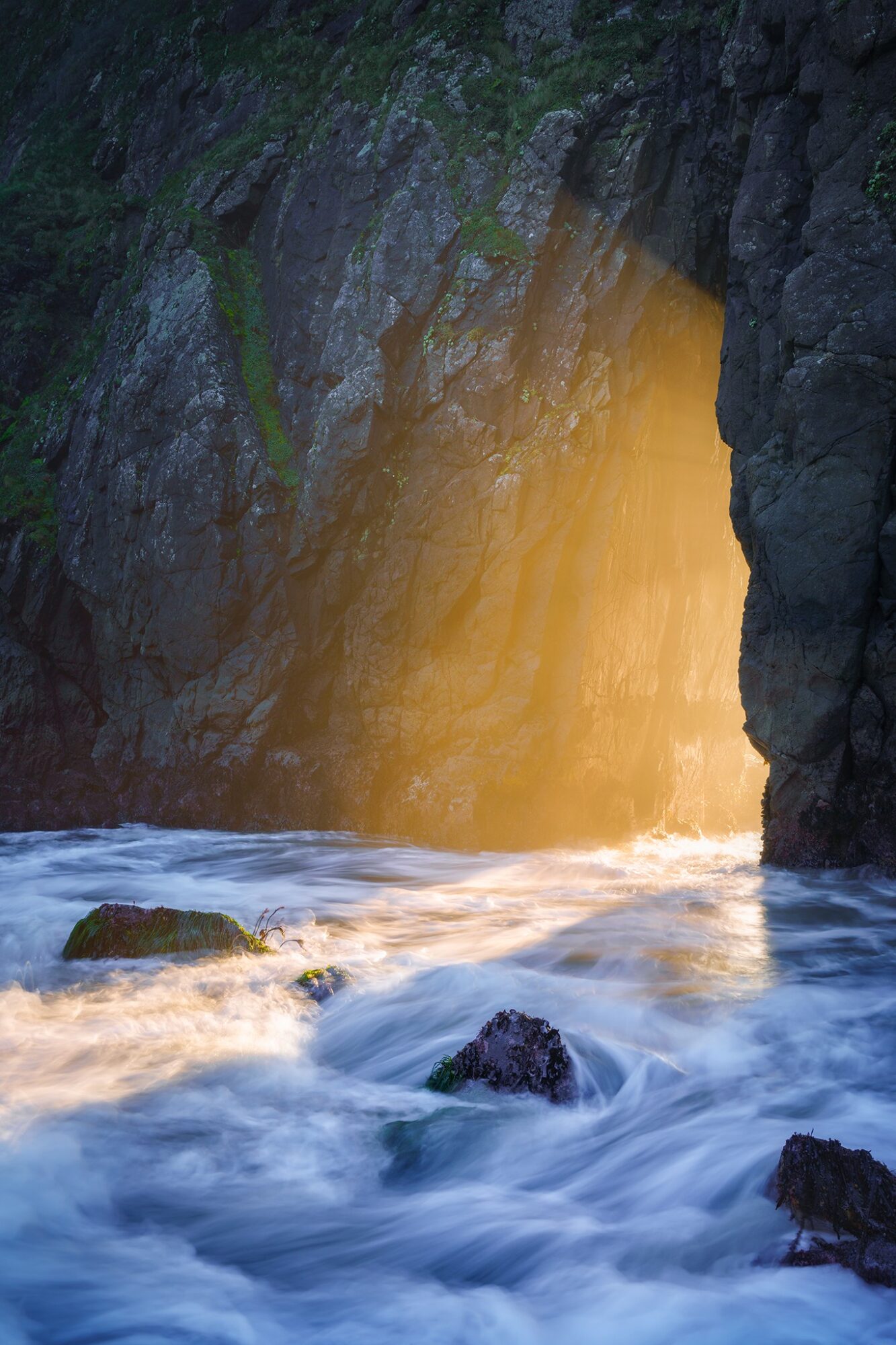 Sunlight streams through a cave opening into the ocean, illuminating water and rocks below.