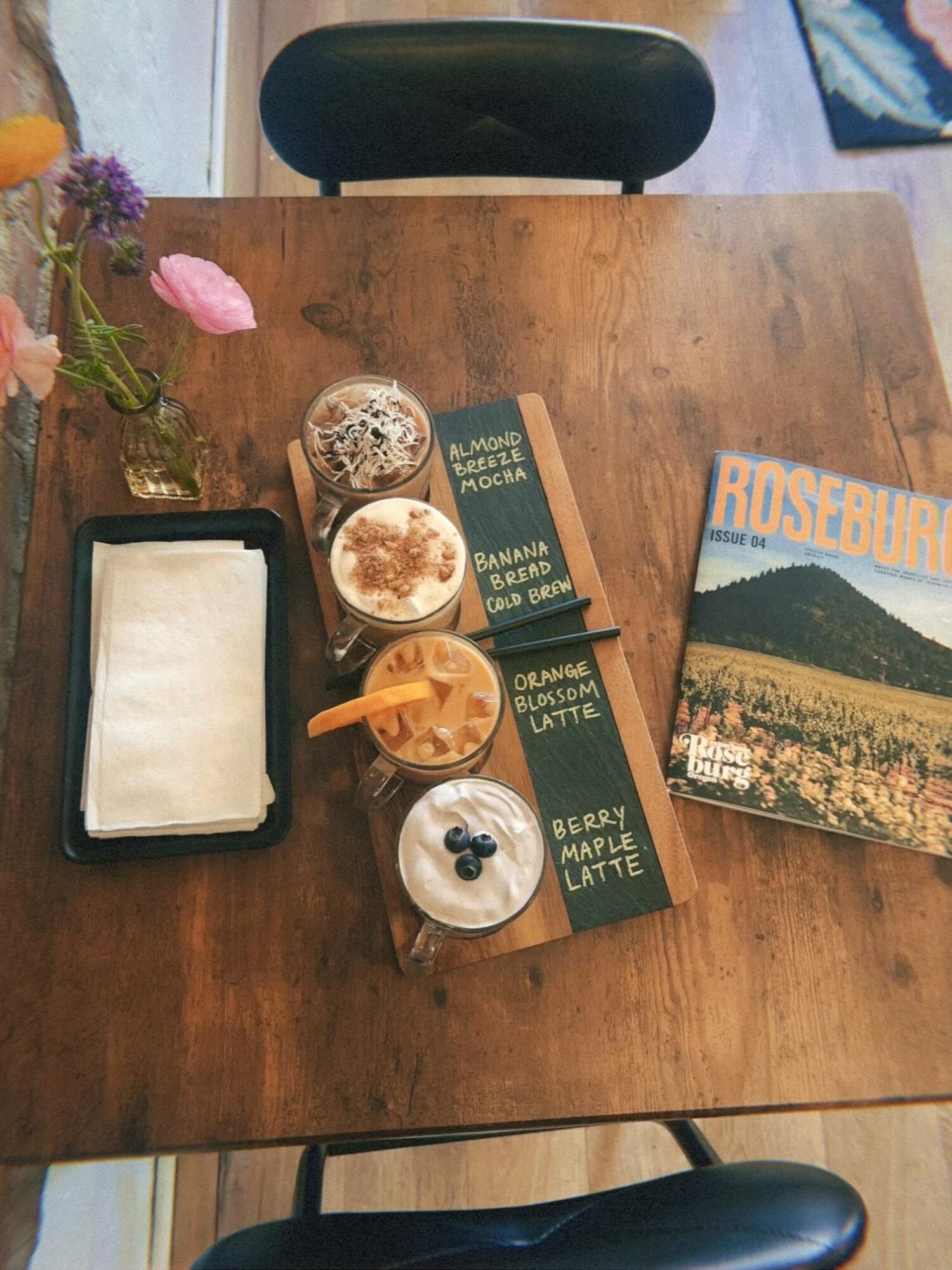 Wooden table with flowers, ice cream, a book, and a magazine, viewed from above.