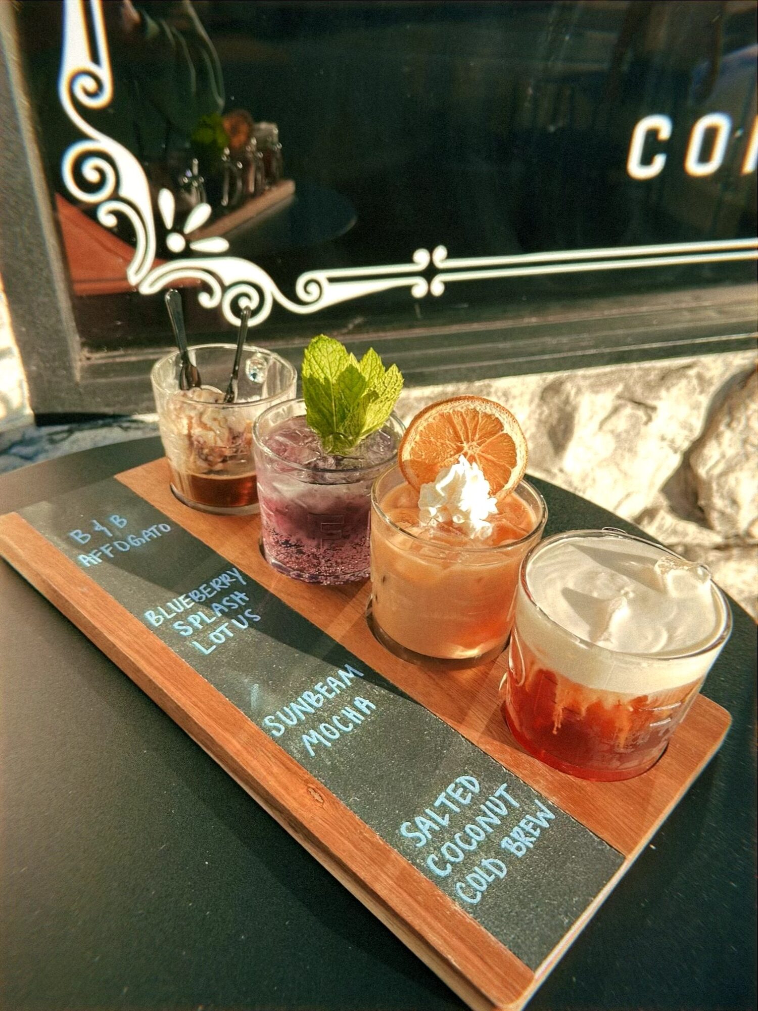 Four colorful drinks with garnishes on a wooden board, with a blackboard menu in the background.