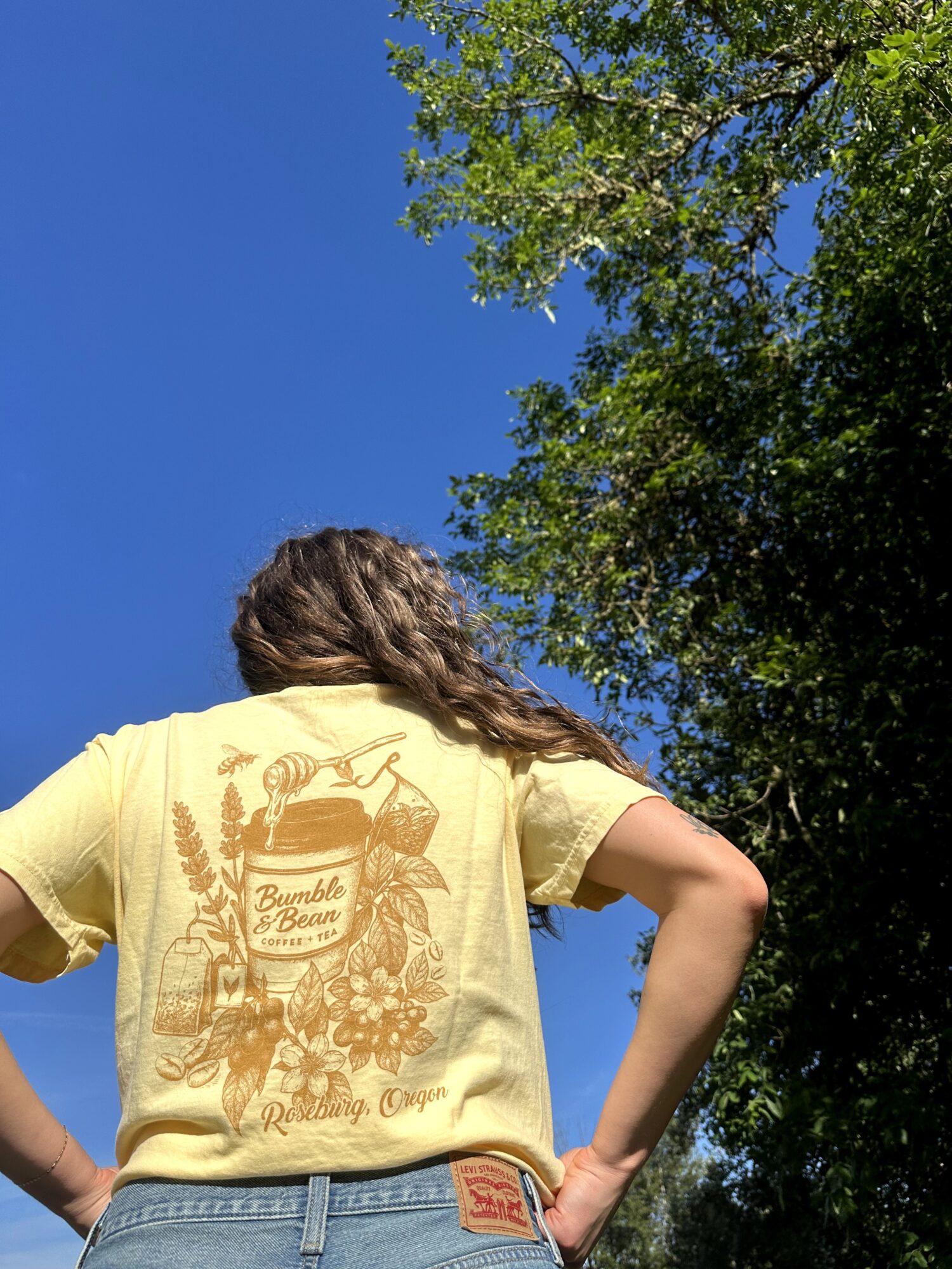 Person with long hair wearing a yellow T-shirt with a graphic design, standing outdoors with trees and blue sky.