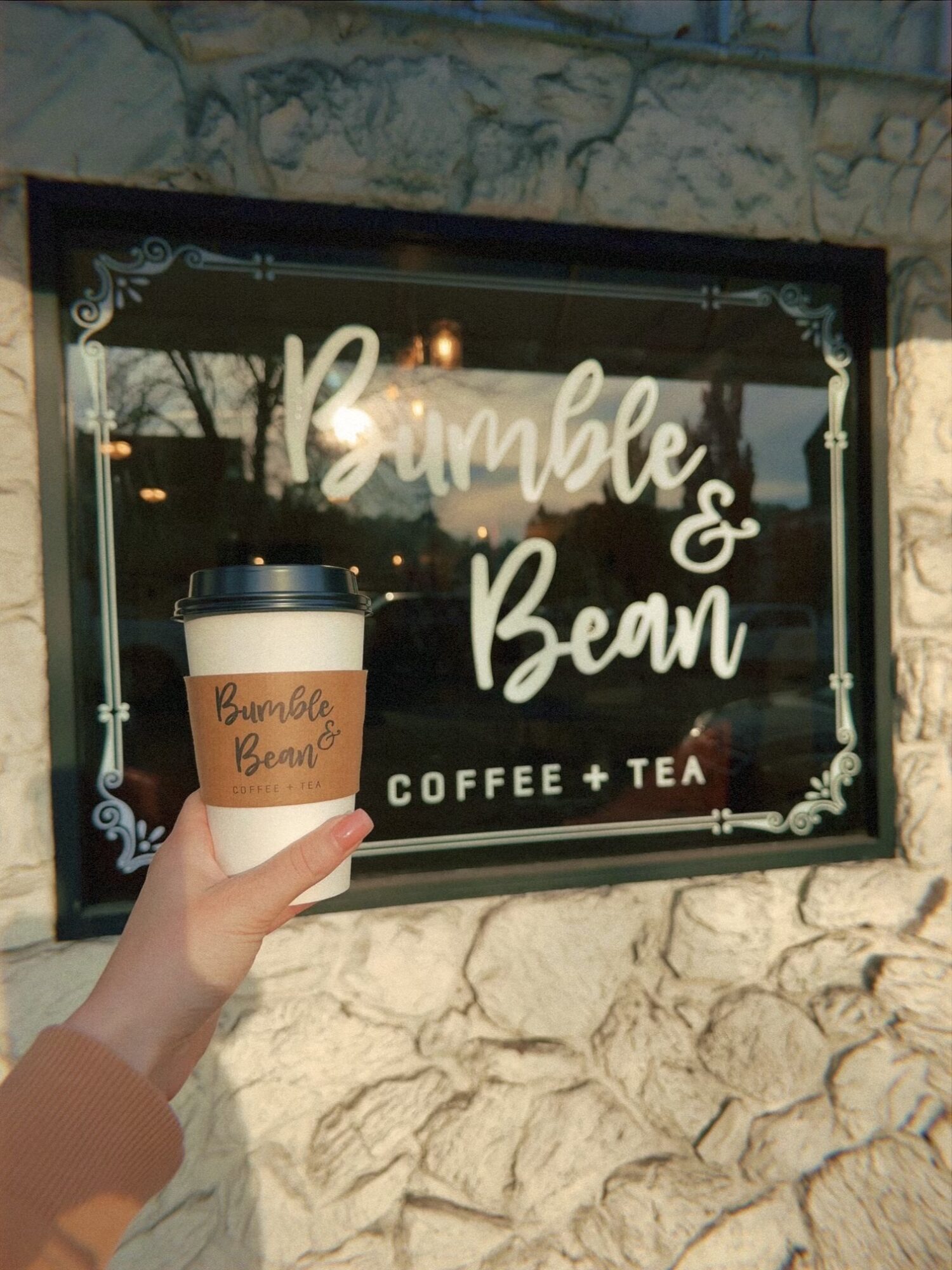 Person holding a coffee cup in front of a sign for Bumble & Bean coffee and tea shop, with a stone wall background.