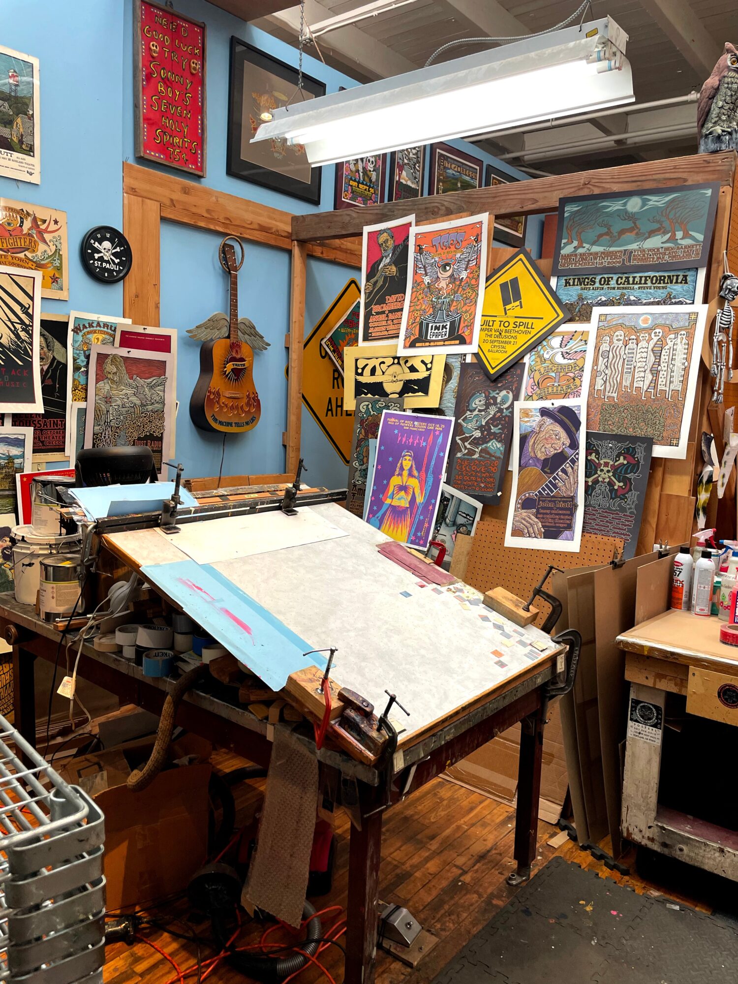 Art studio workspace with a large table, art supplies, and colorful artwork on the walls and bulletin board.