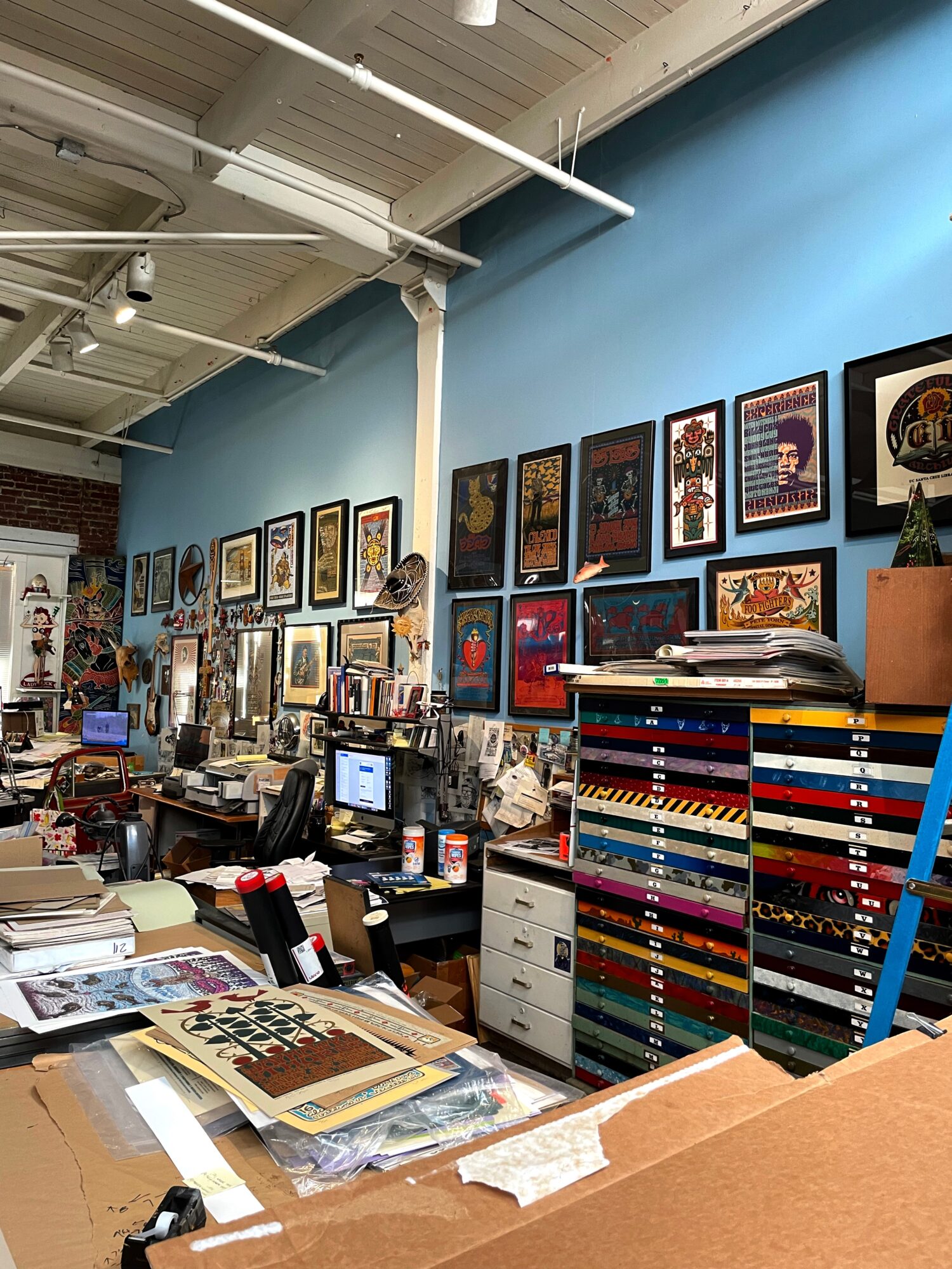 Art studio with framed artwork on blue wall, desks, shelves, and colorful storage drawers, cluttered with papers and art supplies.