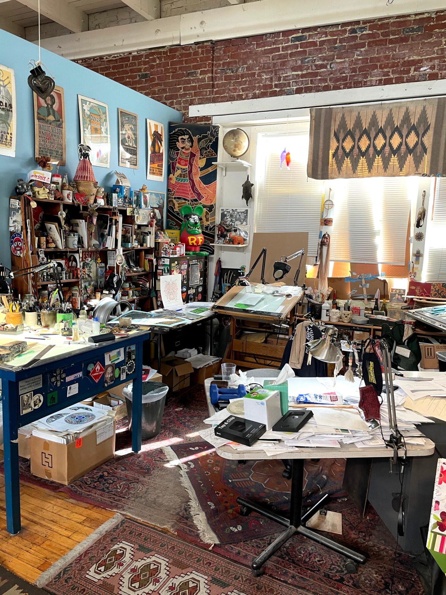 Cluttered room with desks, shelves, artwork, and various objects, including boxes, papers, and decorations, in a creative workspace.