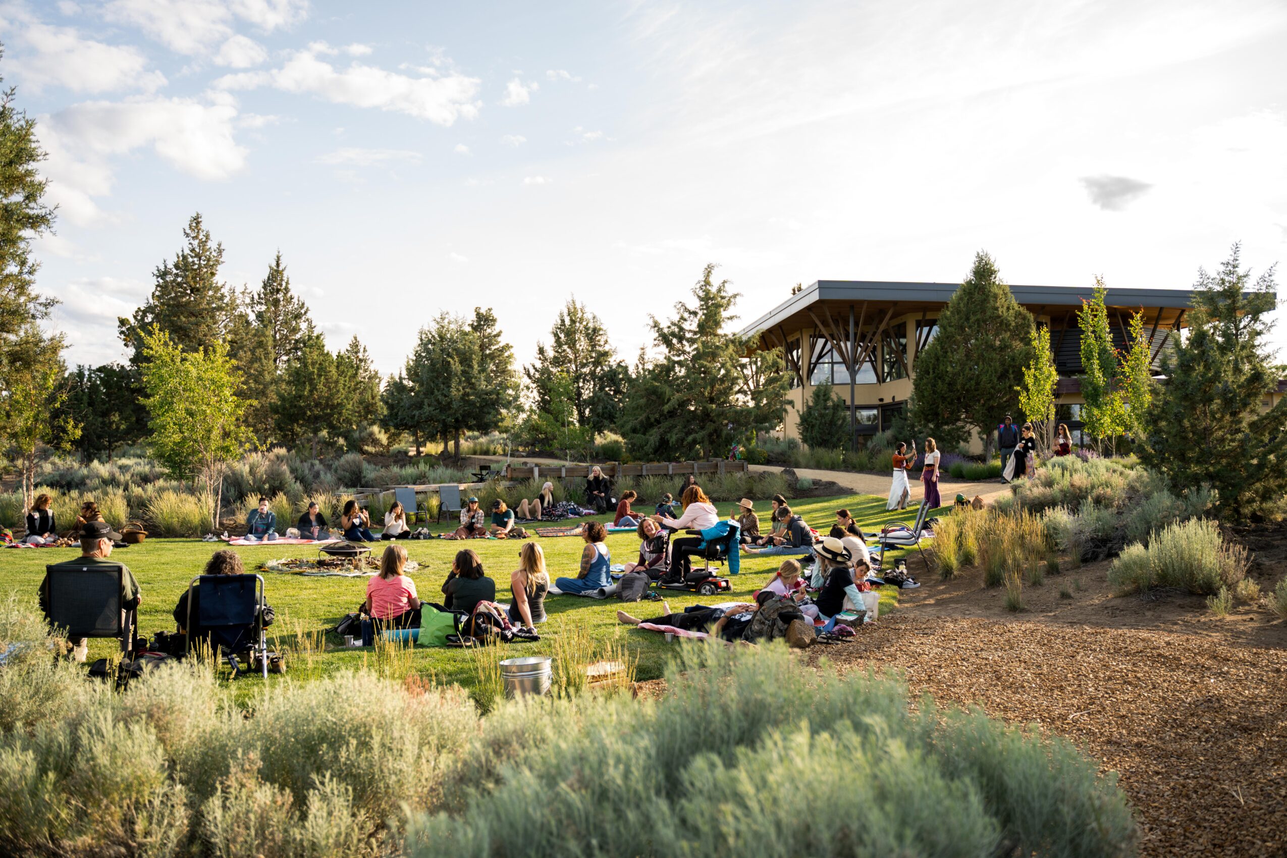 People sitting on chairs and blankets in a park with trees and a building in the background.