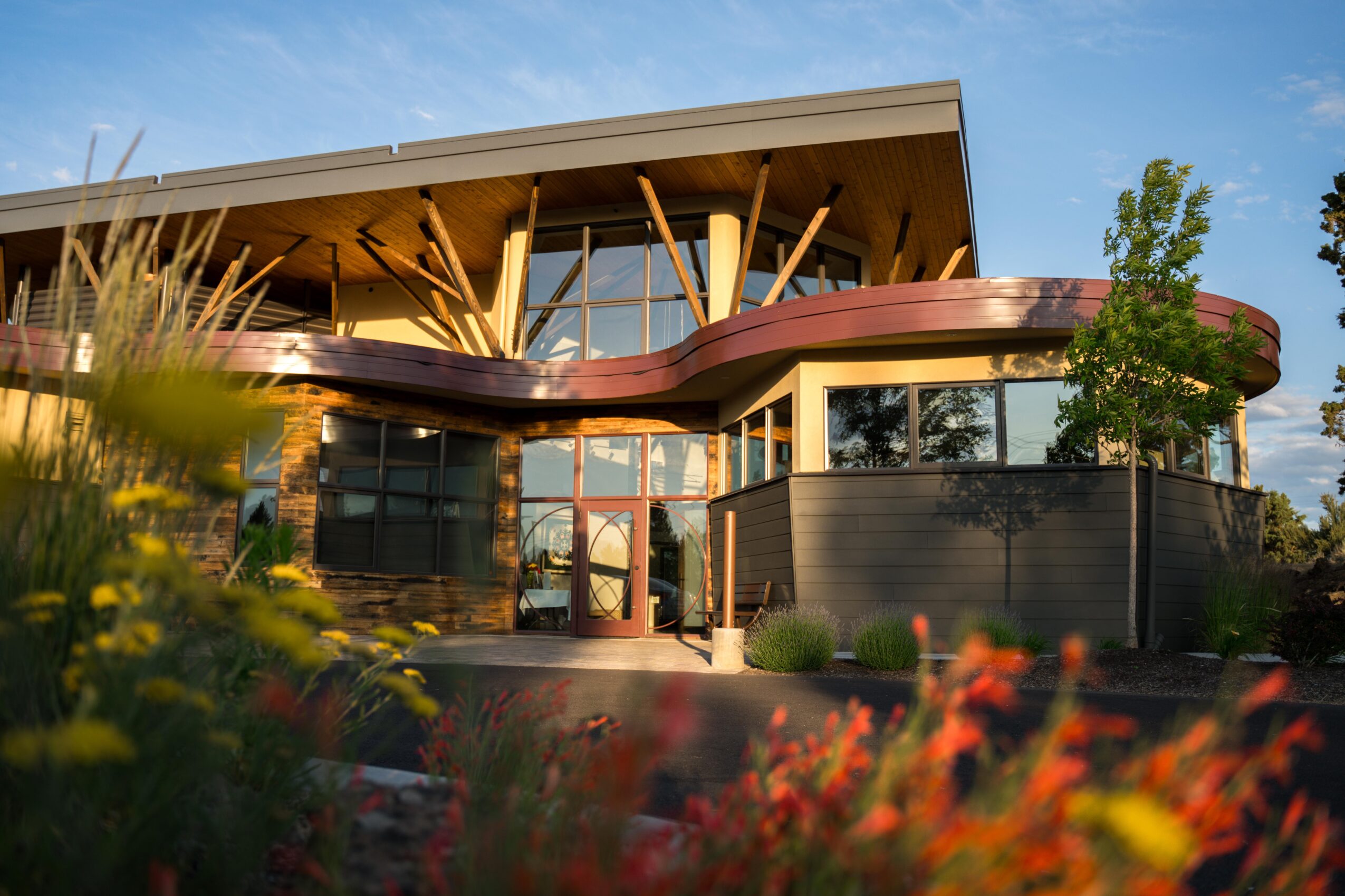 Modern building with large glass windows, wooden beams, and a curved balcony, surrounded by plants and flowers.