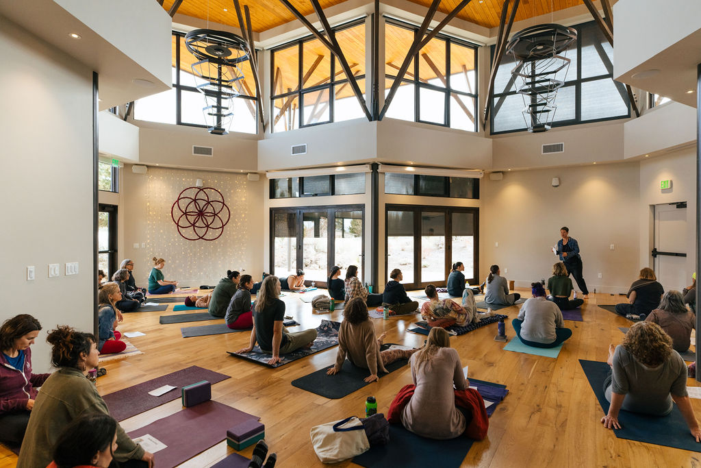 People practicing yoga or meditation in a spacious, well-lit room with large windows and wooden floors.
