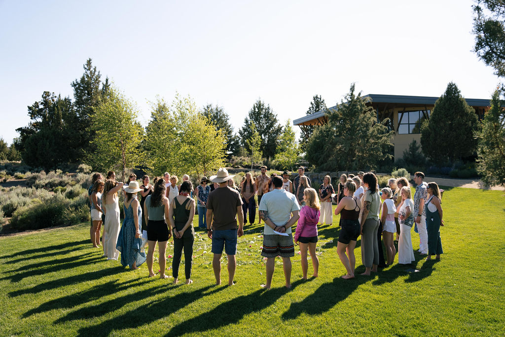 Group of people gathered outdoors on a grassy area with trees and a building in the background.