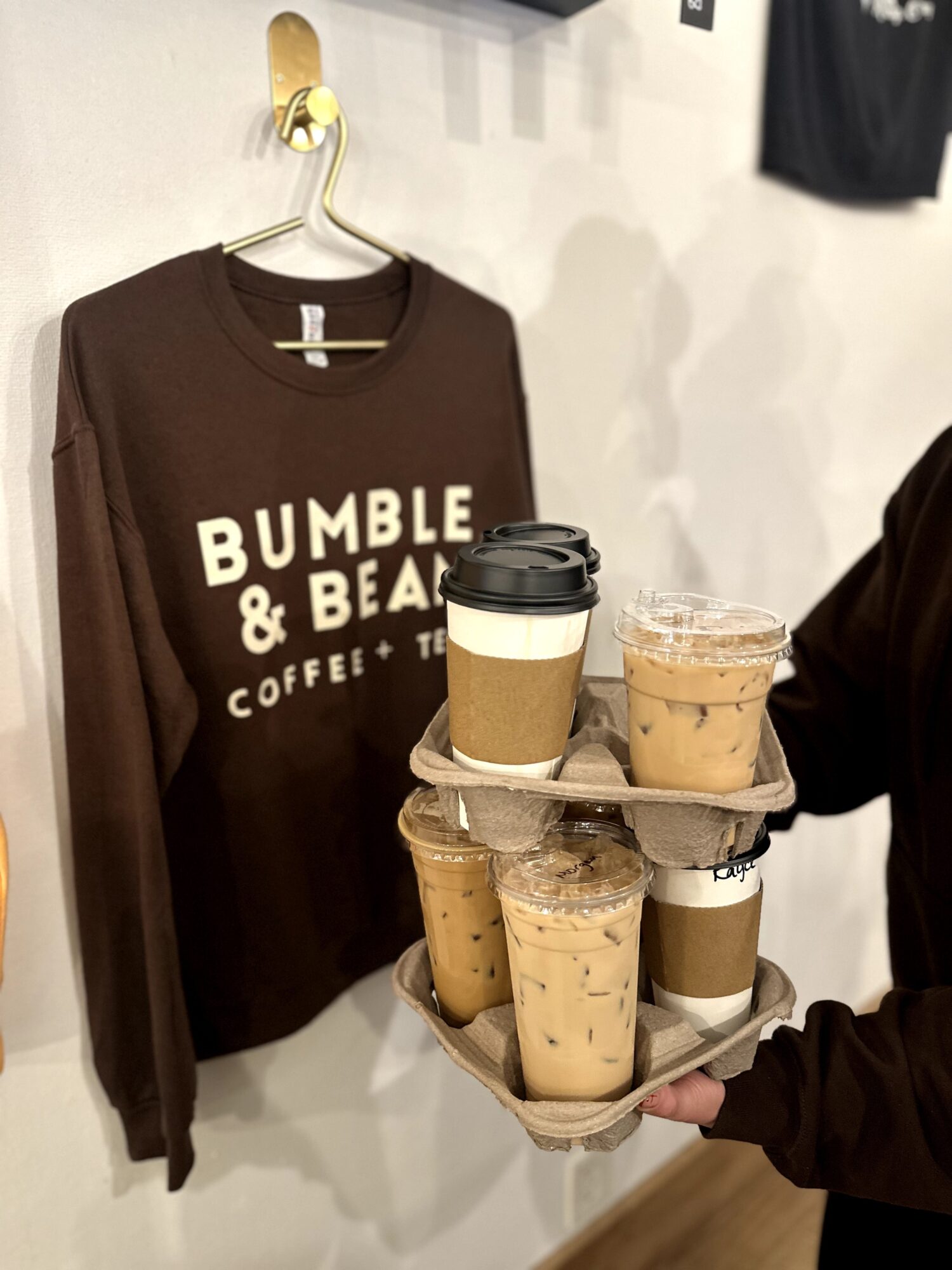 Brown sweatshirt with white text hangs on wall, person holds tray with six iced coffee drinks, background includes a black shirt.