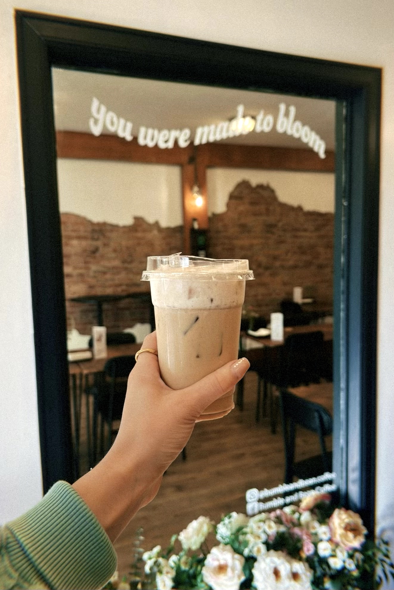 Person holding a glass of iced coffee or latte in front of a mirror with a sign that reads 'You were meant to bloom.'