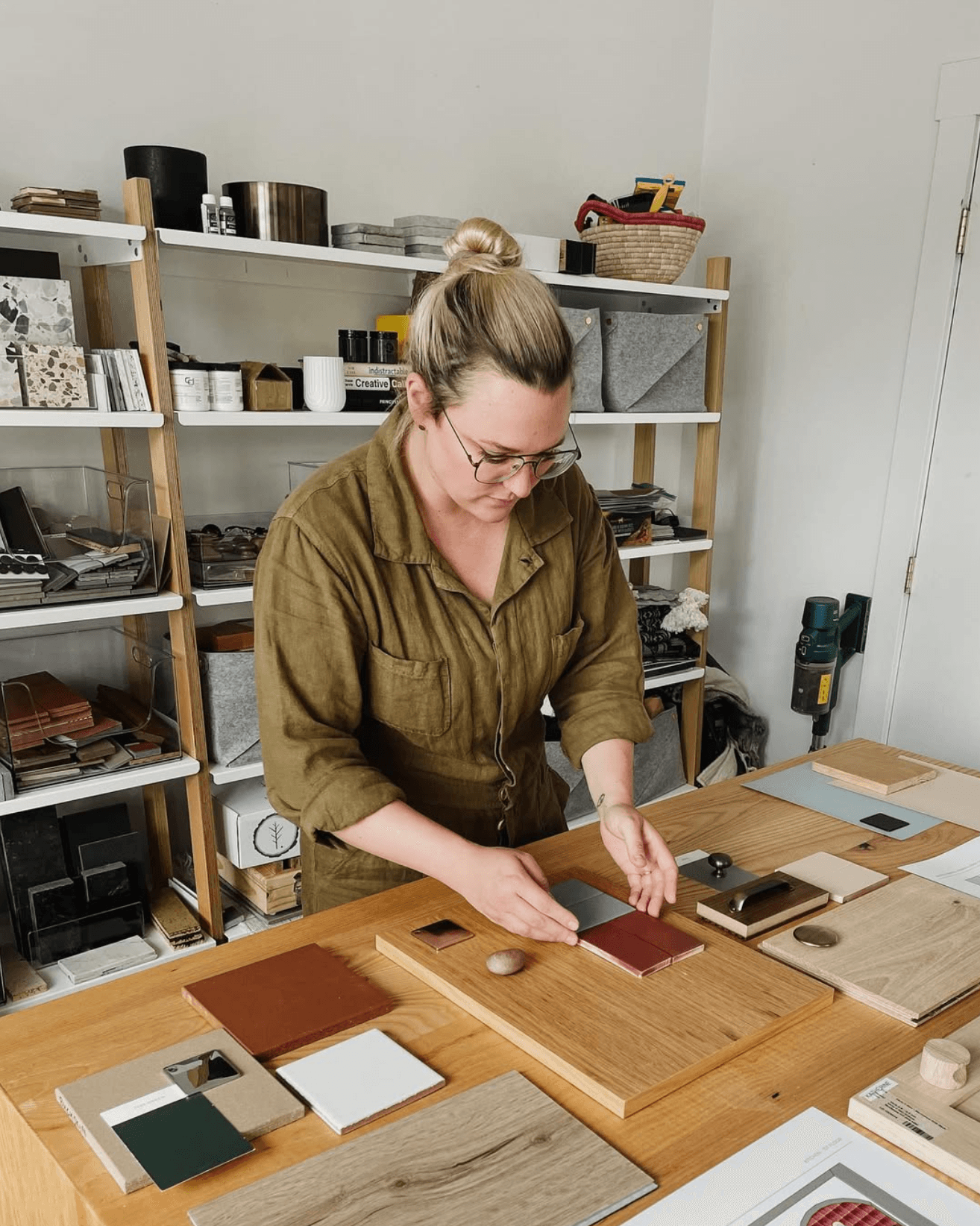 Woman with glasses and blonde hair in a bun, wearing a green shirt, working at a table with wood samples and swatches in a workshop.
